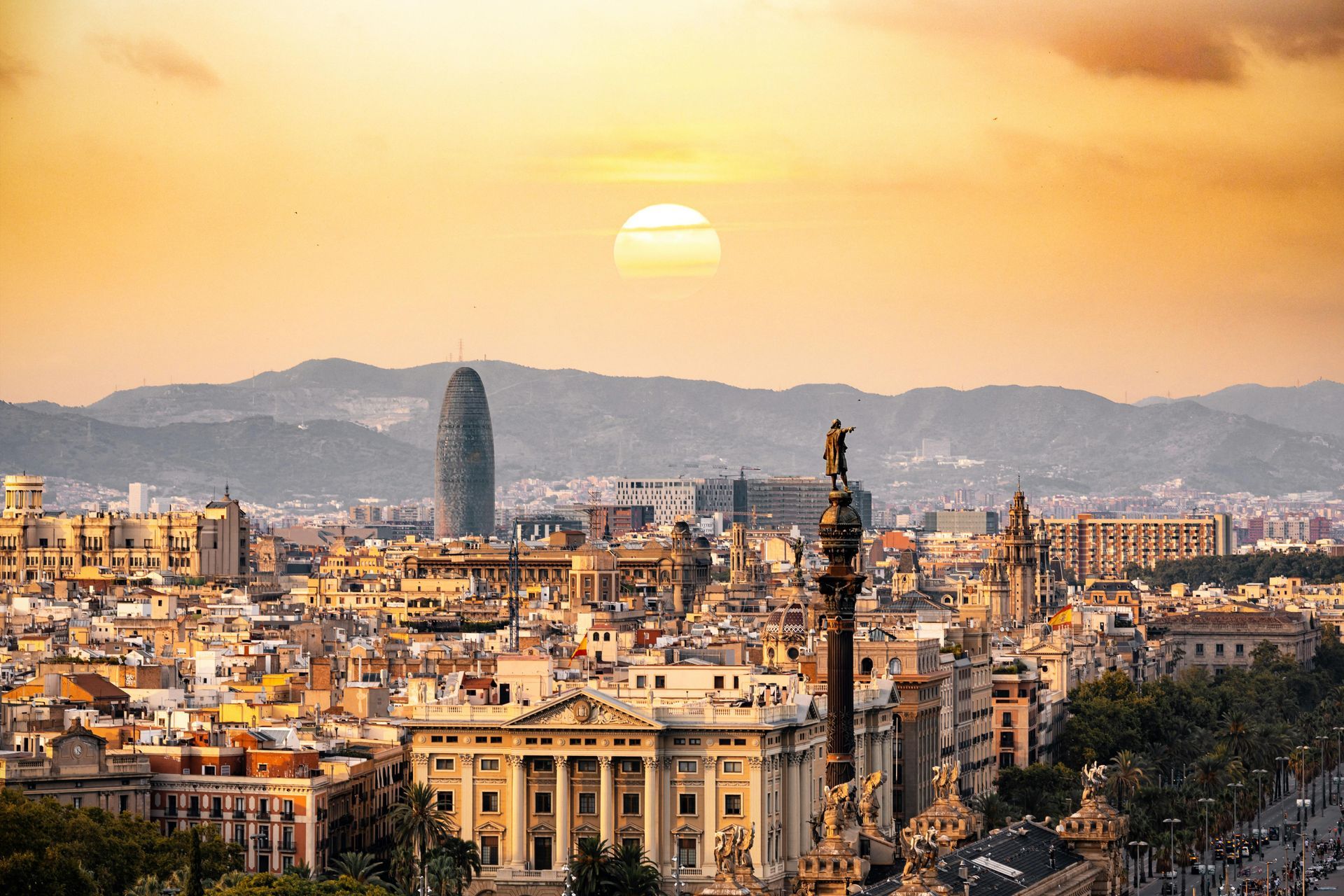 The sun is setting over a city in Spain with a statue in the foreground.