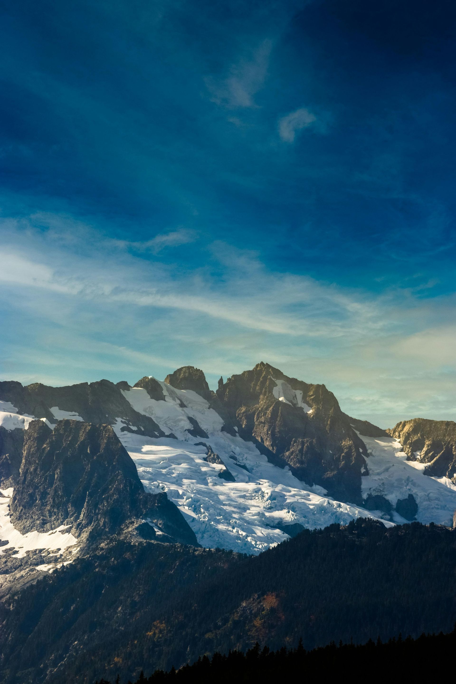 Mountain range with patches of snow and a blue sky. Dark forest in the foreground.