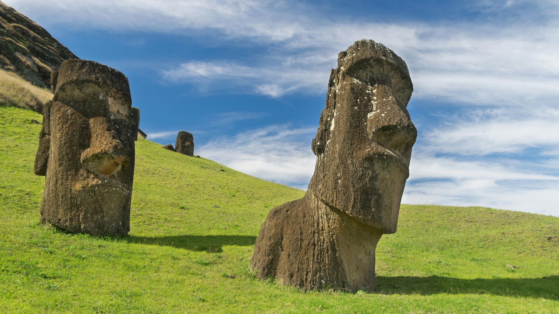 Moai statues on a green grassy hillside under a partly cloudy blue sky.