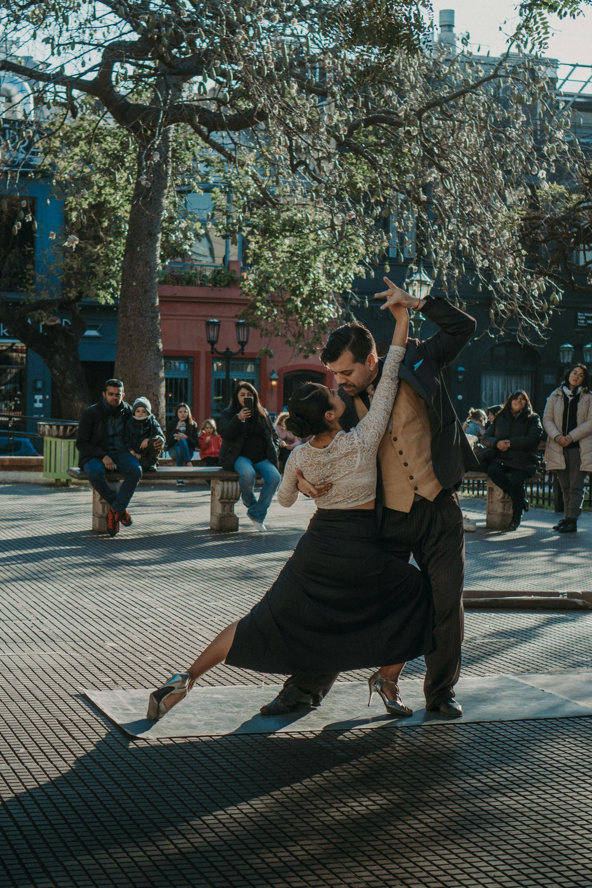 A couple dances the tango outdoors in a plaza. The woman, in a black skirt, leans back dramatically. The man wears a suit.