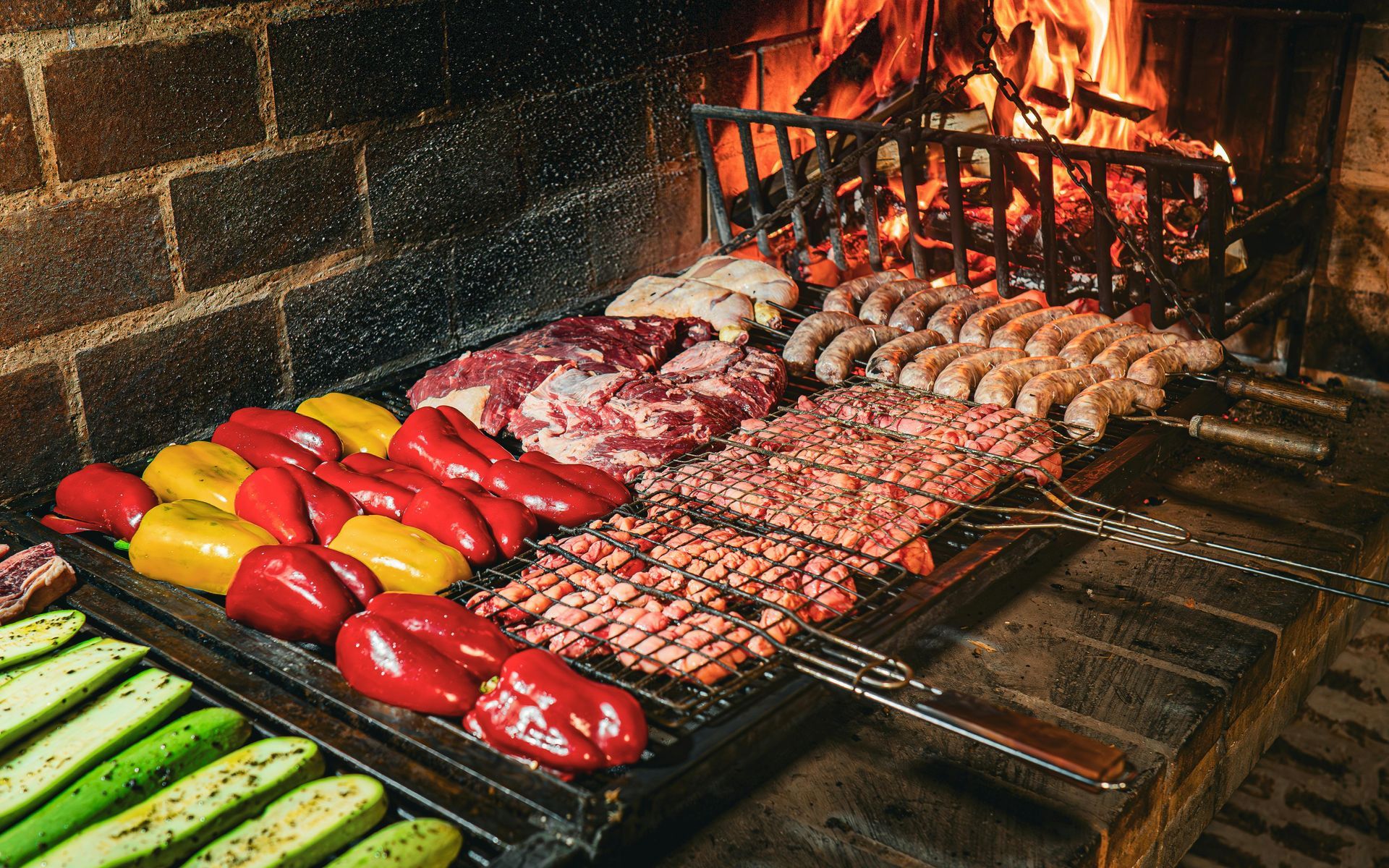 Grilled meat and vegetables on a bbq with a fire blazing in the background. Includes peppers, zucchini, steak, and sausages.