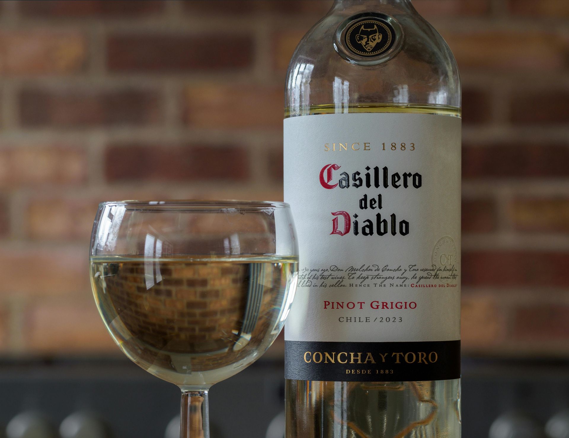 A glass of white wine and a bottle of Casillero del Diablo Pinot Grigio sit in front of a brick wall.