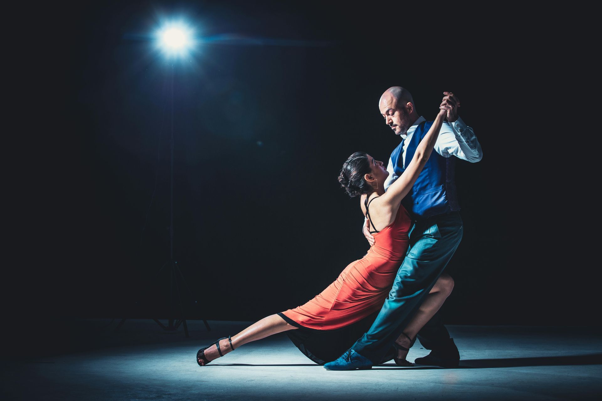 Man and woman in tango pose; man in blue vest, woman in red dress; dancing in a dark setting lit by a spotlight.