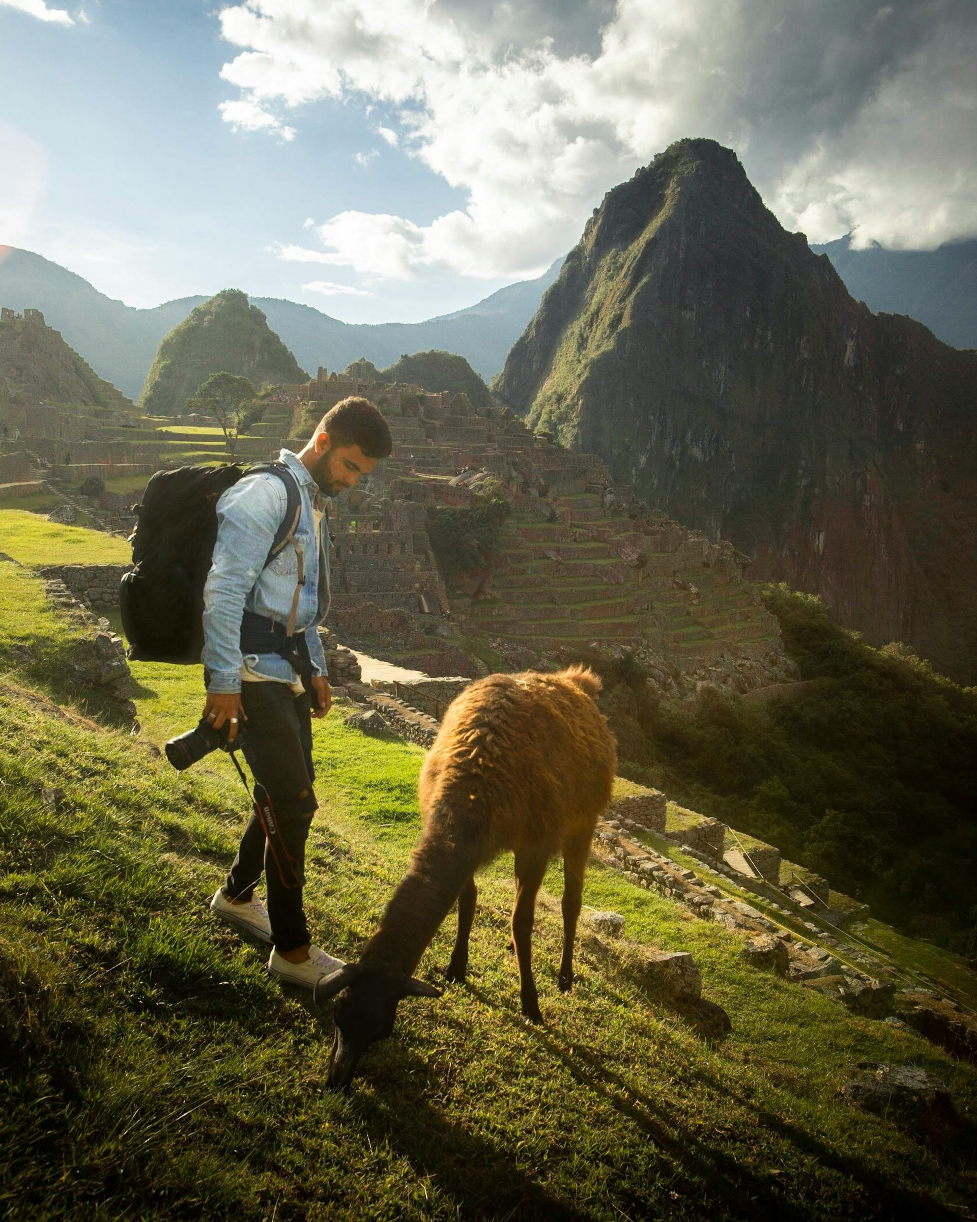 Man with backpack and camera standing next to a llama on a grassy slope overlooking Machu Picchu ruins.