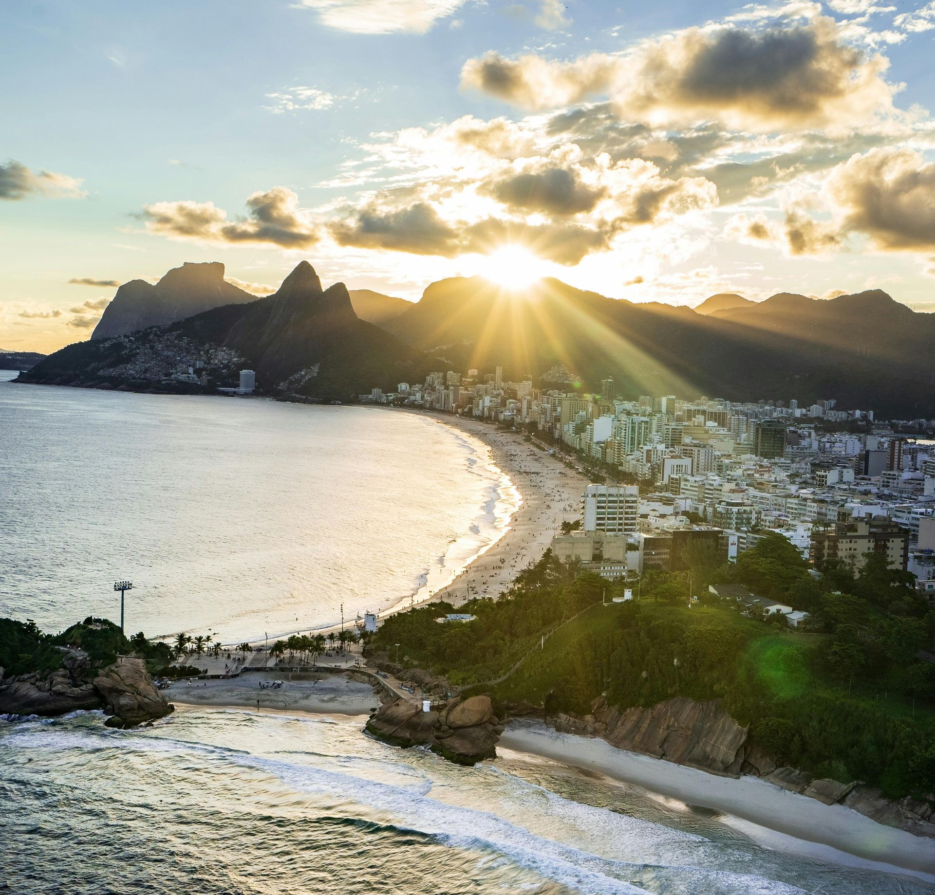 Beach at sunset in Rio de Janeiro, Brazil. Sun bursts over mountains, illuminating the coastline, beach, and city.