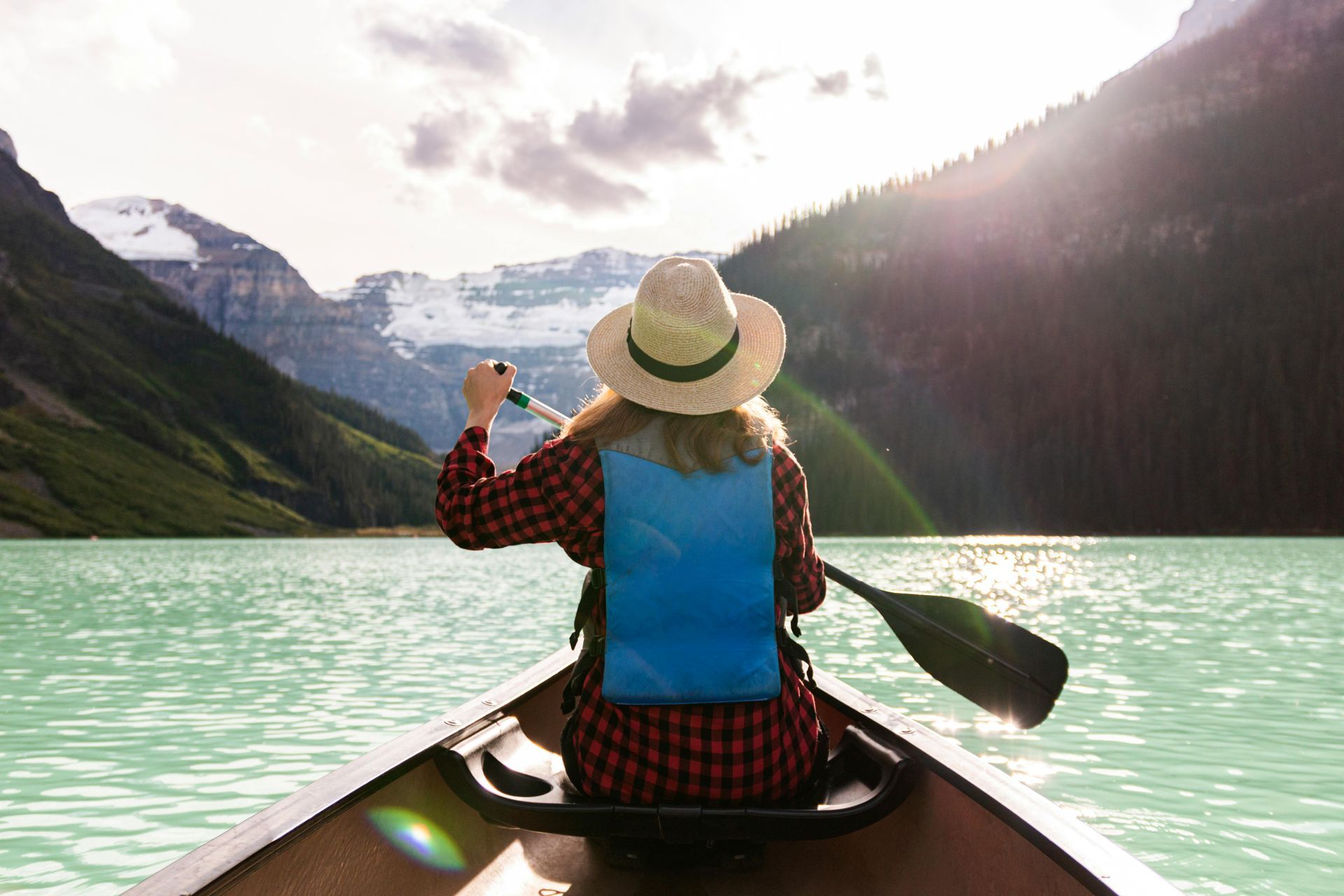 Woman canoeing on a turquoise lake, mountains in the background, wearing a hat and red plaid shirt.