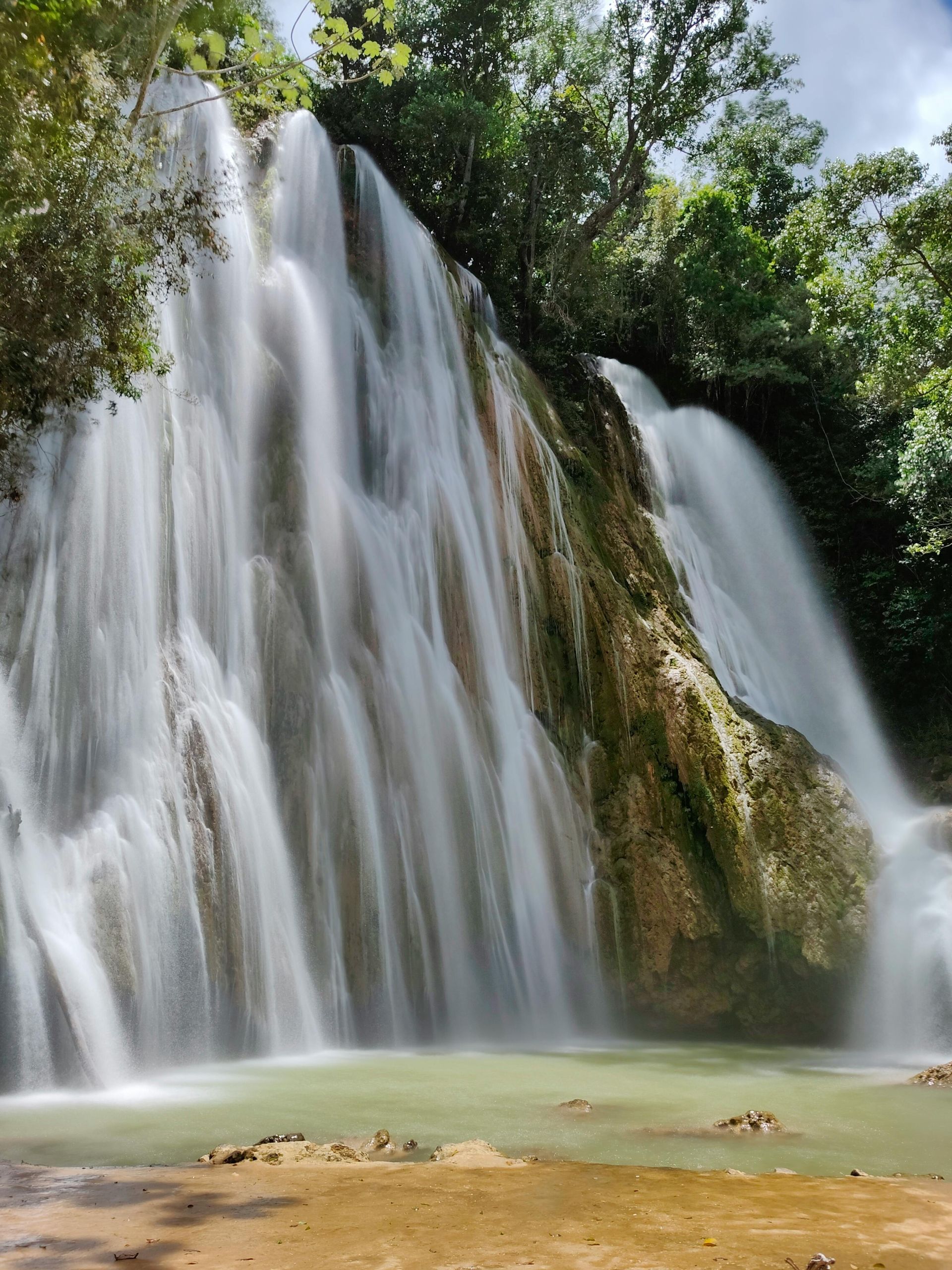 A cascading waterfall rushes down a rocky cliff into a light green pool, surrounded by lush green foliage.