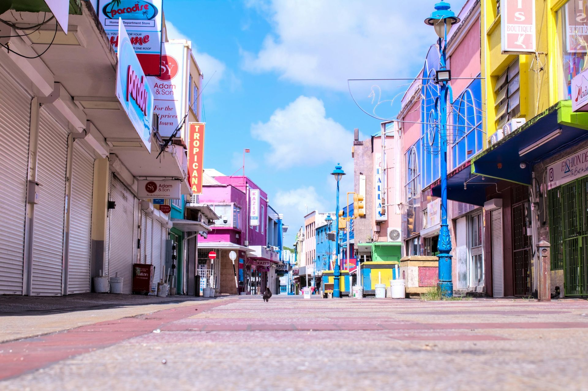 A colorful street with shops closed, pink, yellow, and blue buildings. Clear blue sky with a few clouds.