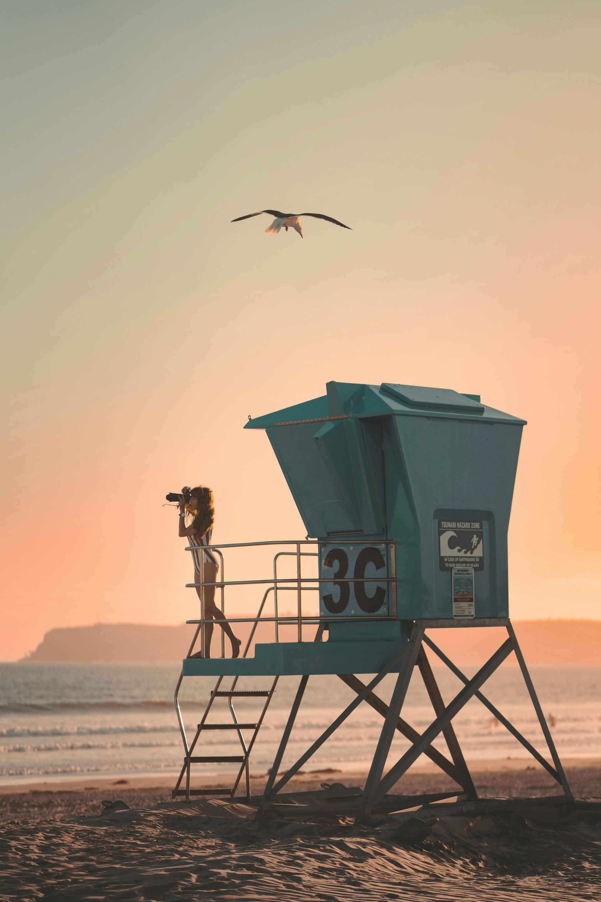 A lifeguard tower on a beach at sunset, a woman standing and looking through binoculars, a seagull flying overhead.