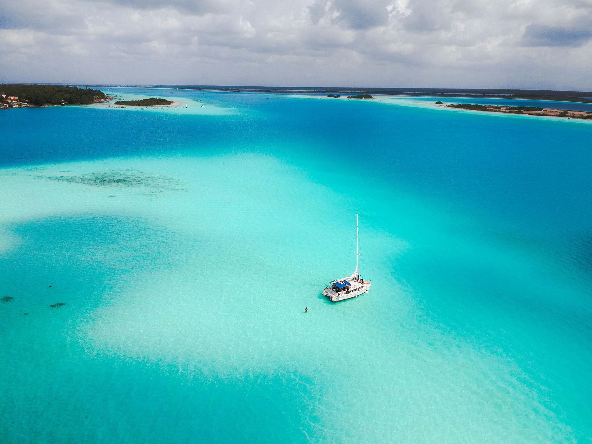 Aerial view of a turquoise lagoon with a sailboat. Bright blue water, white boat, and cloudy sky.
