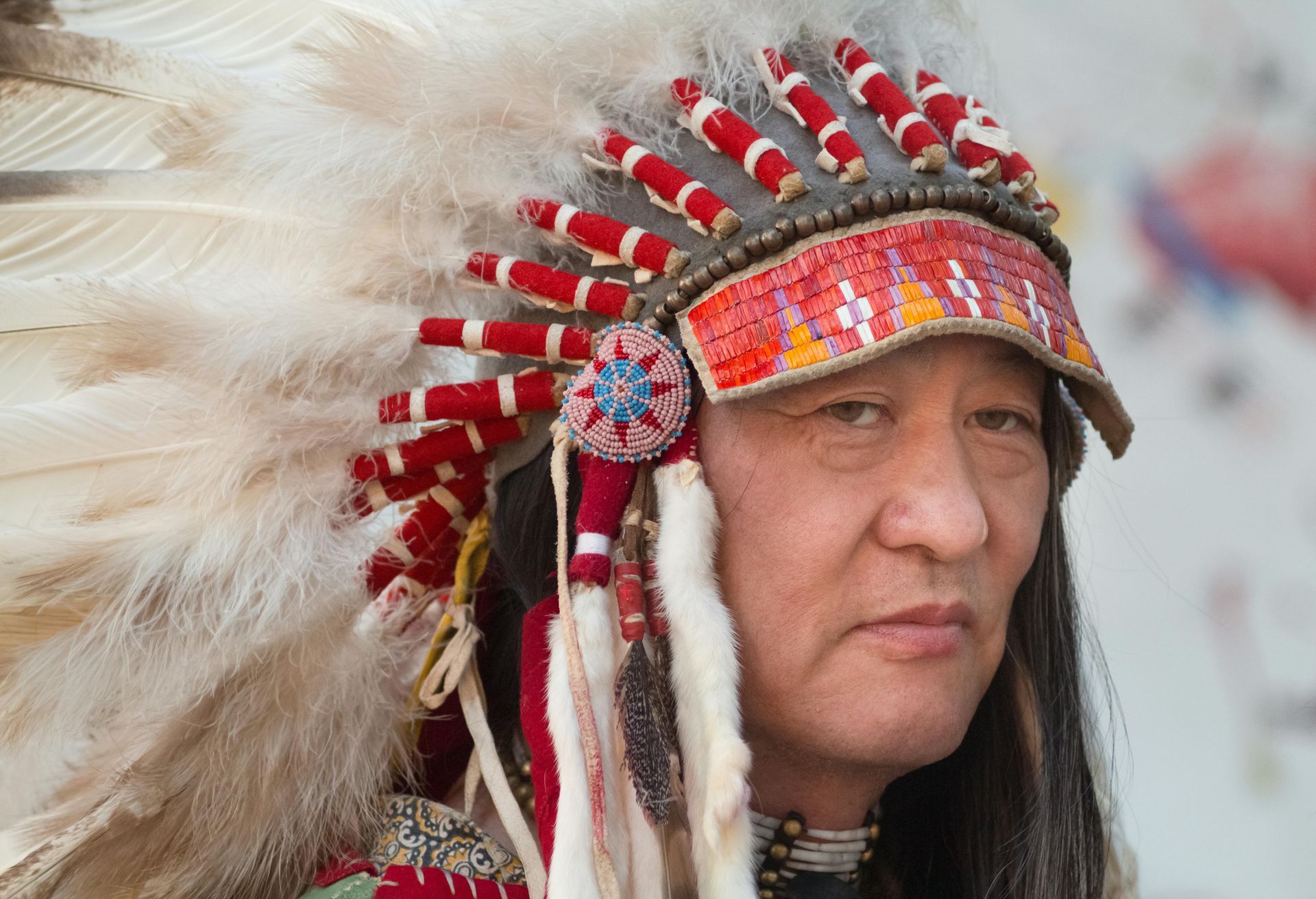 Man wearing a feathered Native American headdress, looking at the camera. Headdress is red, white, and beaded