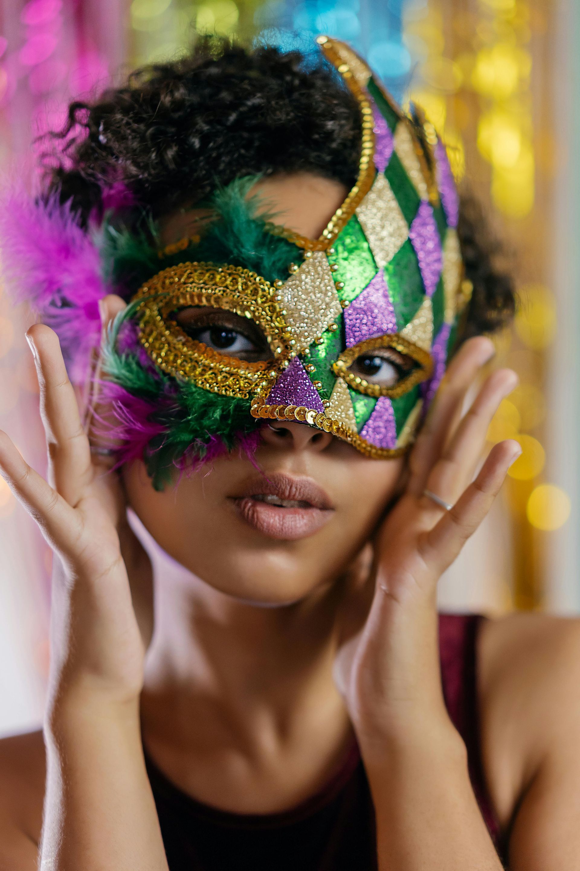 Woman wearing a Mardi Gras mask with green, purple, and gold accents. She is holding her hands up near her face.