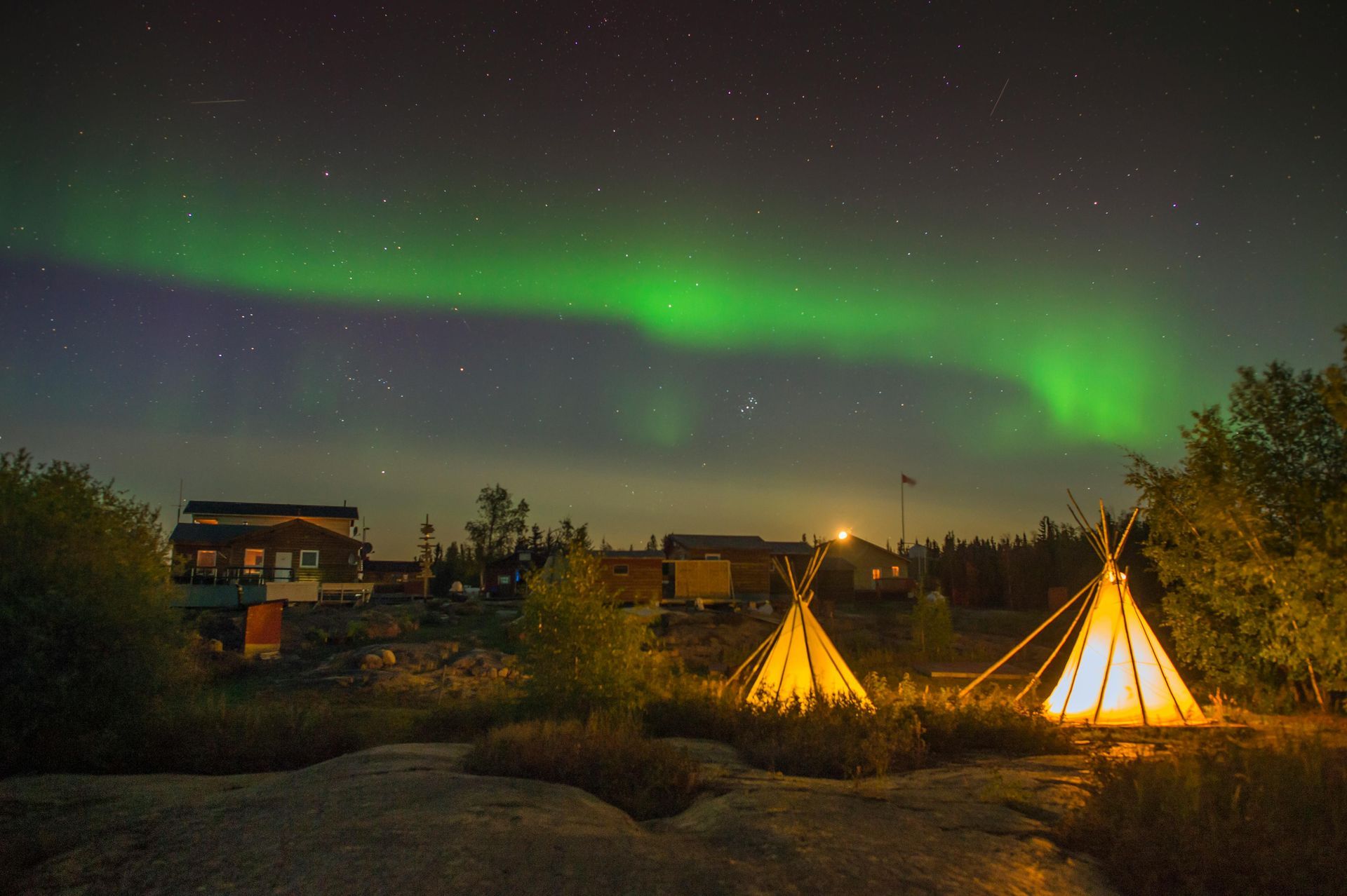 Northern lights illuminate the night sky above a camp with tipis. Green and purple aurora borealis over the sky.