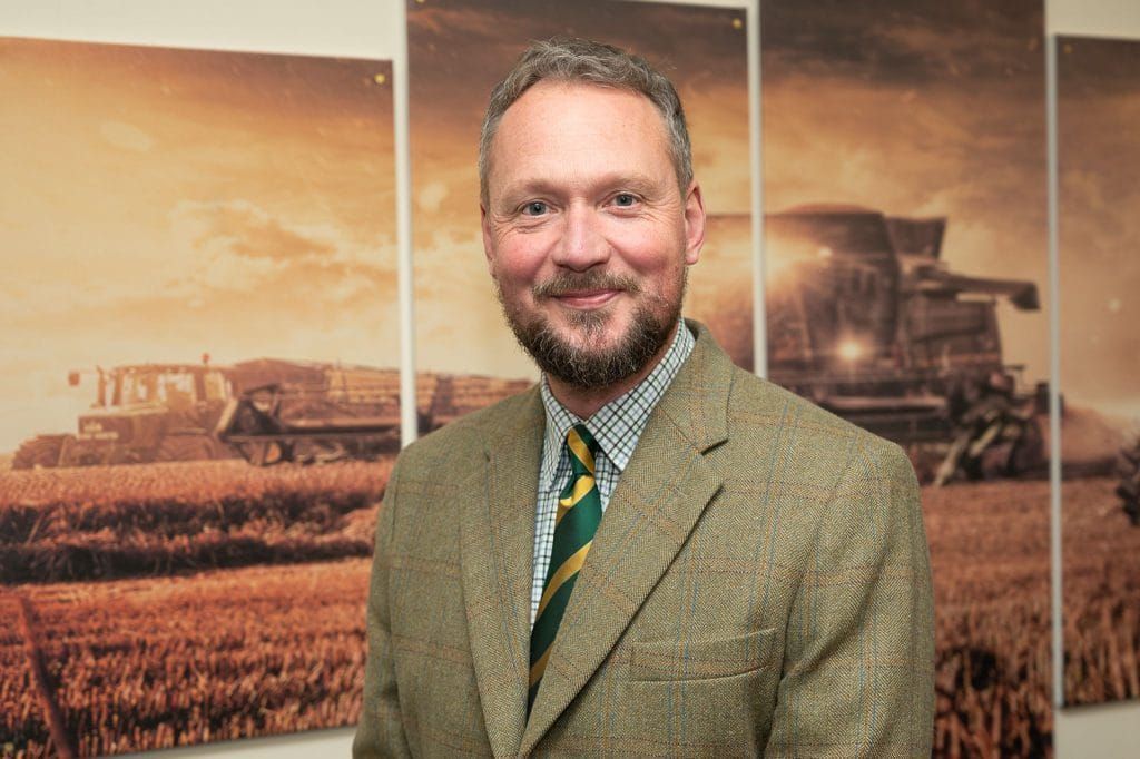 Man in tweed jacket and green tie standing before framed farm landscape prints