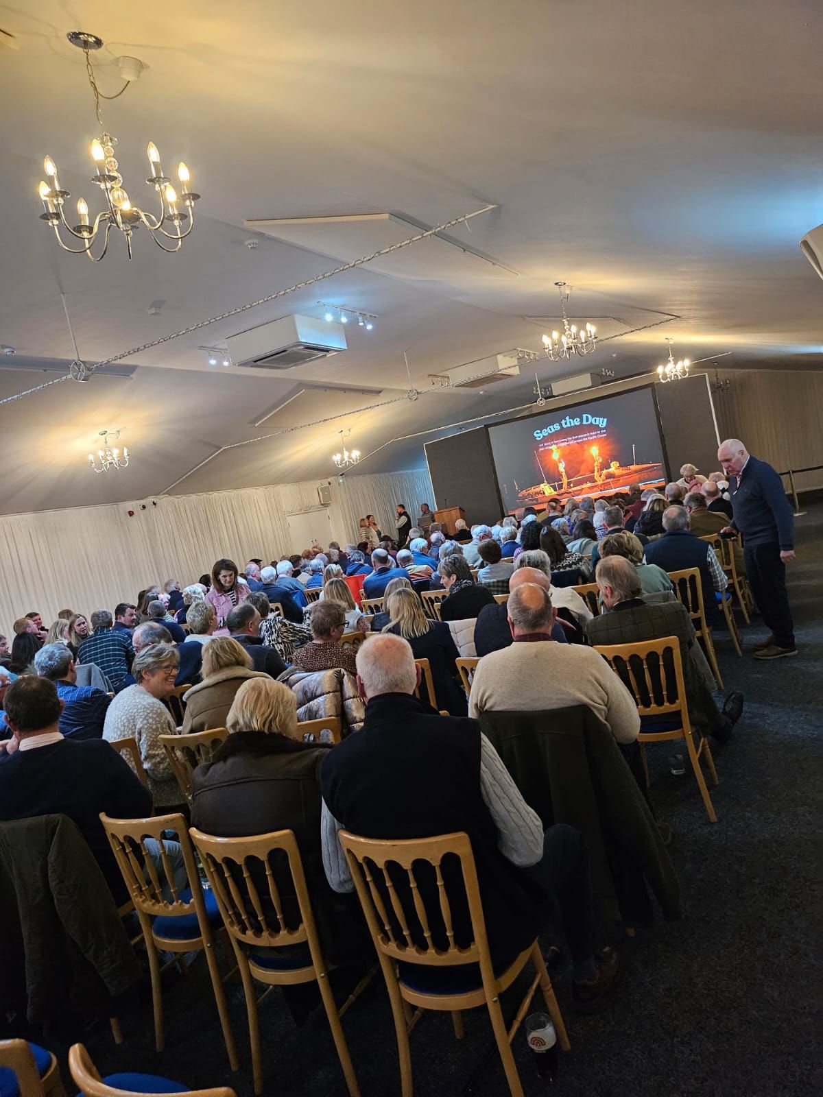 Crowded banquet hall with seated audience facing a lit stage and presentation screen