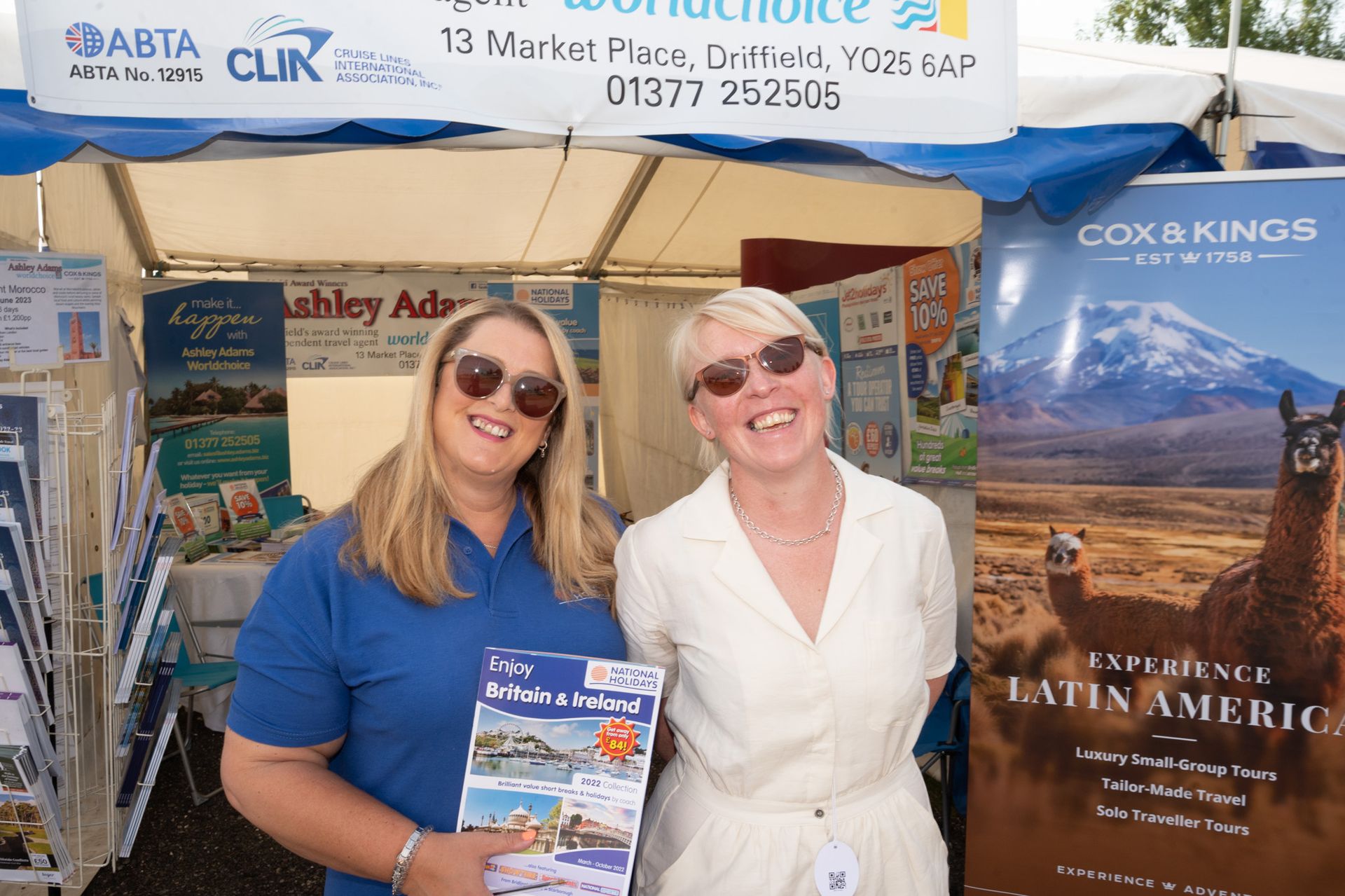 Two smiling people at a travel booth, holding brochures under a canopy, with travel posters behind them