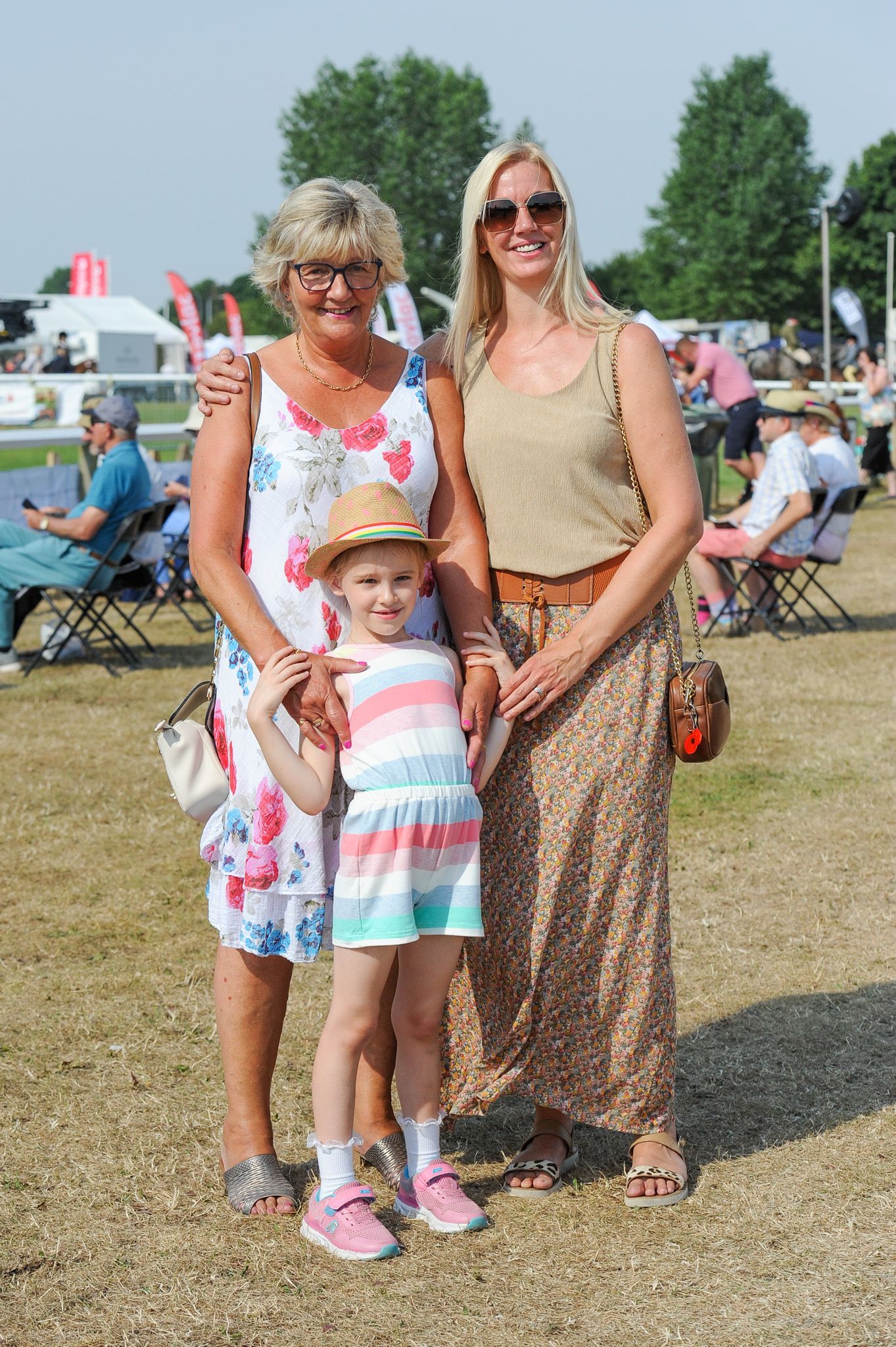 Three people posing outdoors in summer clothing at a fairground, with folding chairs and tents behind them.