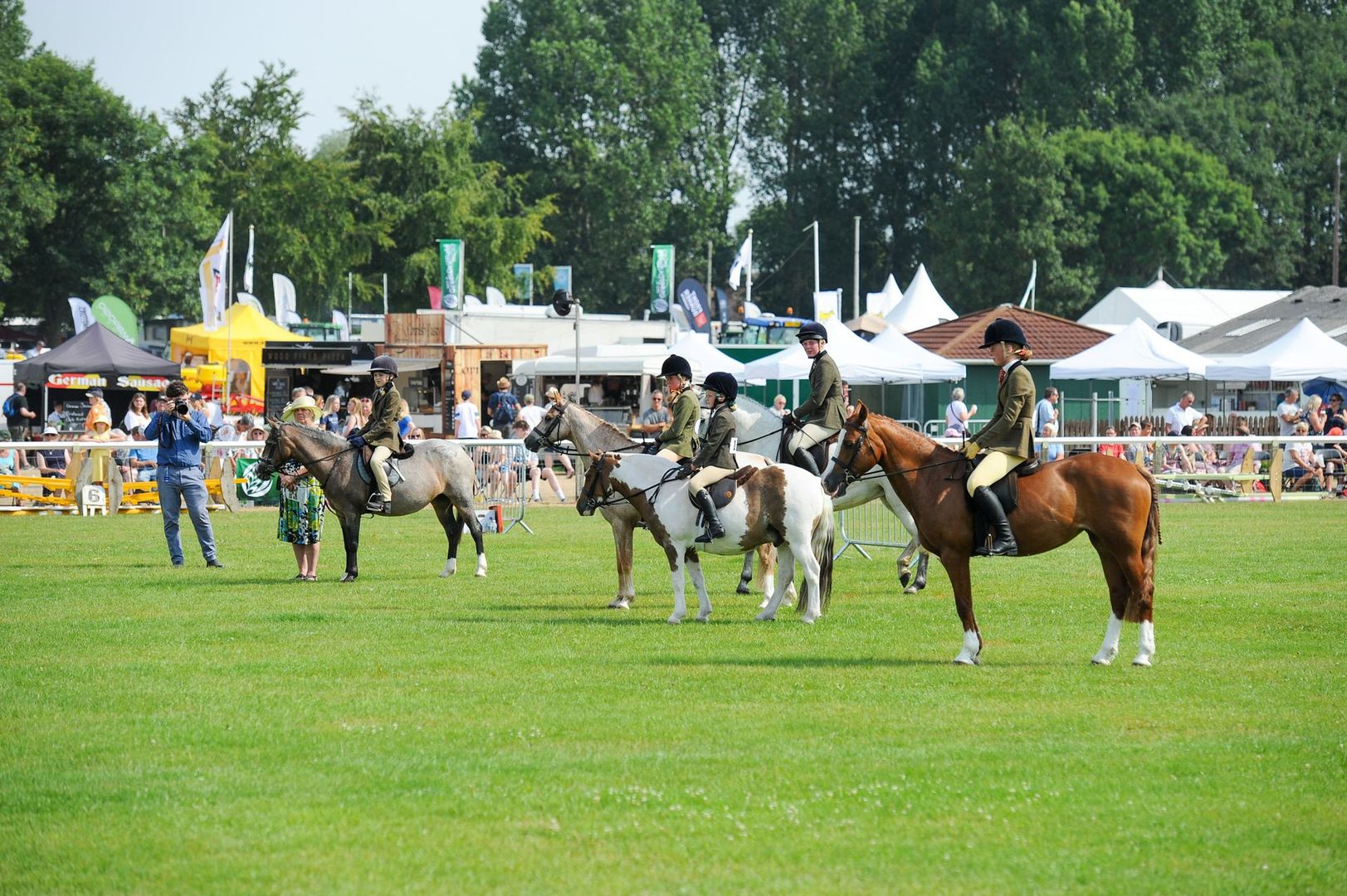 Riders on horses in a grassy show ring with tents and spectators in the background
