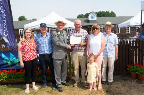 Group posing outdoors at an event, with one man holding a certificate in front of a flower display and tent.