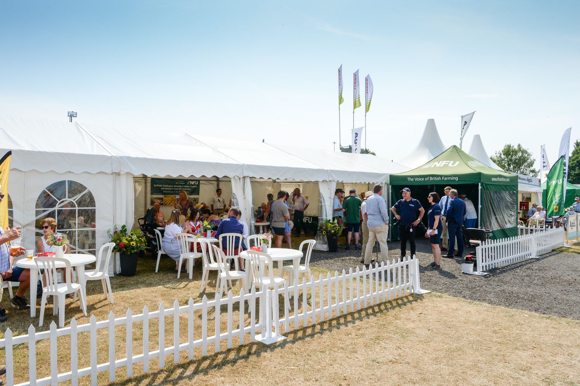 Outdoor event tent with people gathered around tables, white picket fence, and green flags on a sunny day