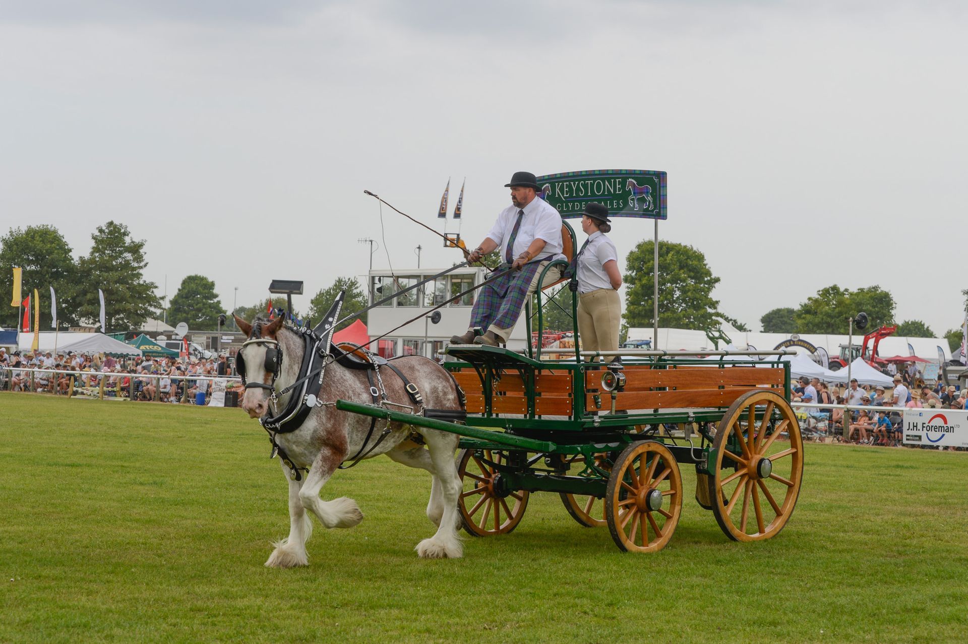 Horse-drawn carriage in a grassy field with two riders, wooden cart, and crowd in the background