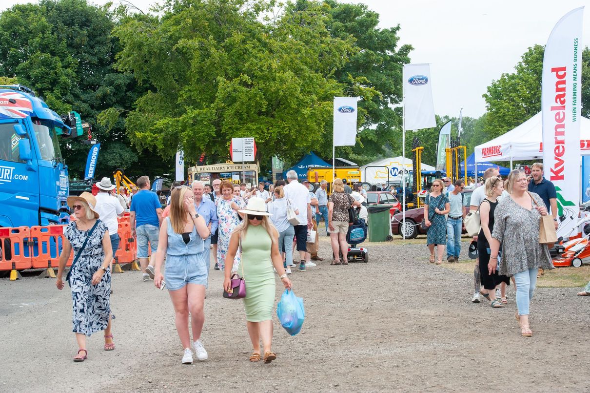 Crowd walking through a festival entrance with flags, tents, and trees on a gravel path