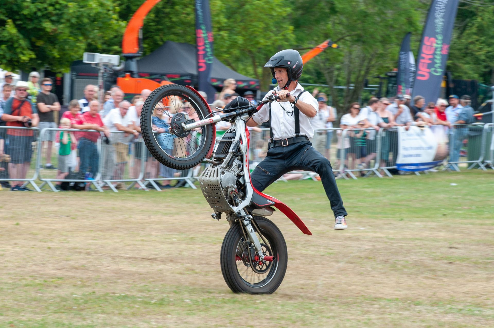 Motorcyclist performing a wheelie at an outdoor event, with a crowd watching behind barriers.
