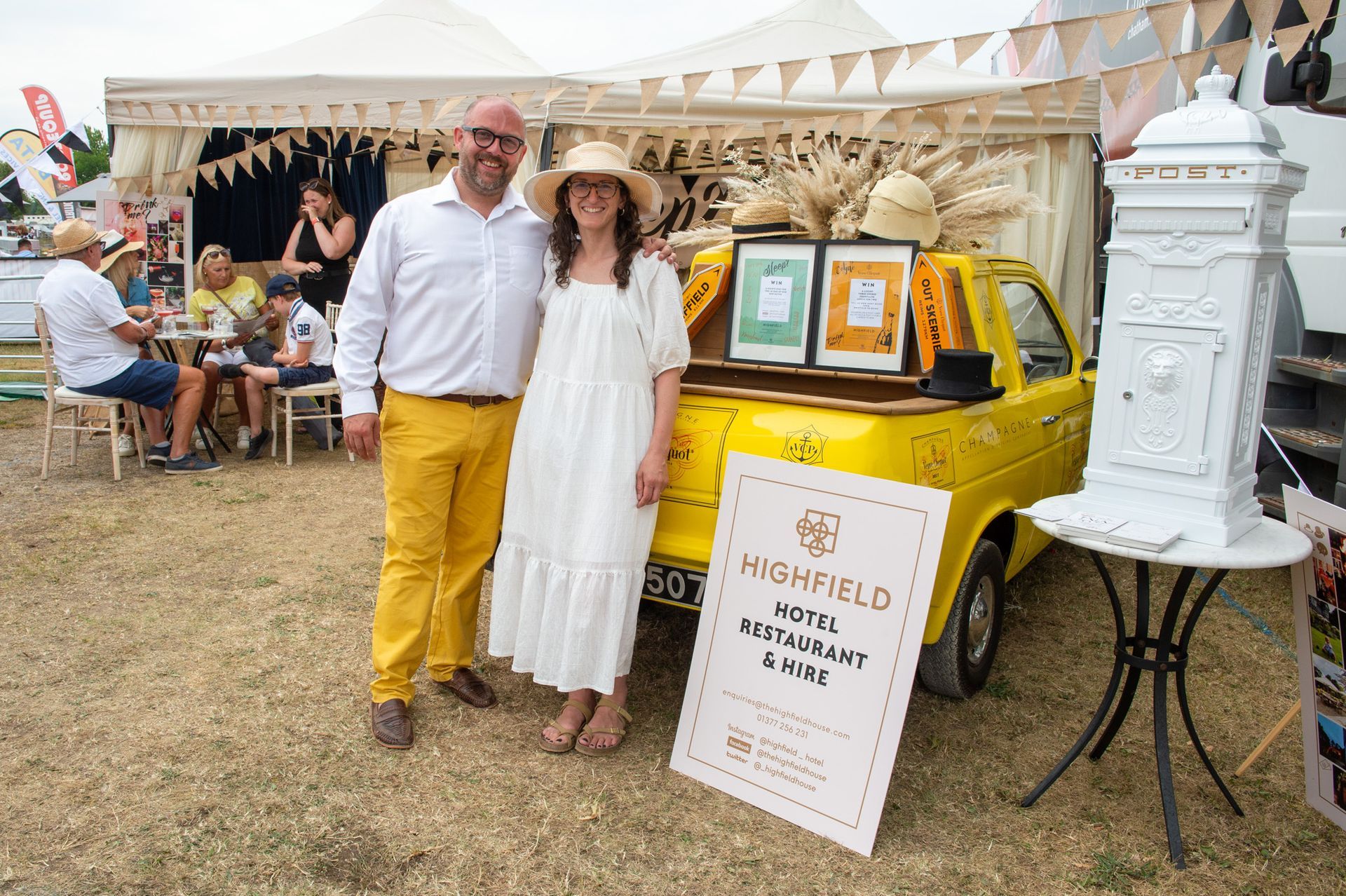 Two people stand by a yellow trailer at an outdoor market display of jars and signs.