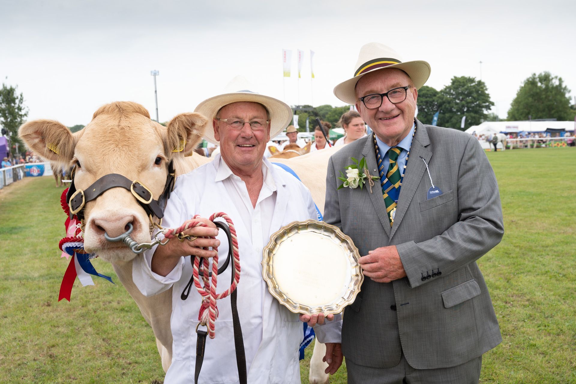 Two men in hats posing with a cream-colored cow at an outdoor fair, one holding a rosette and the other a plaque