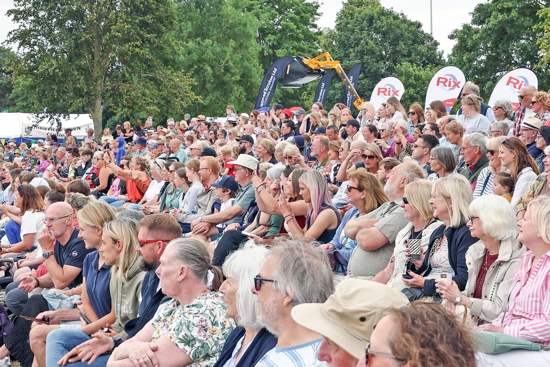 Crowd seated outdoors at a public event, with people holding signs and trees in the background