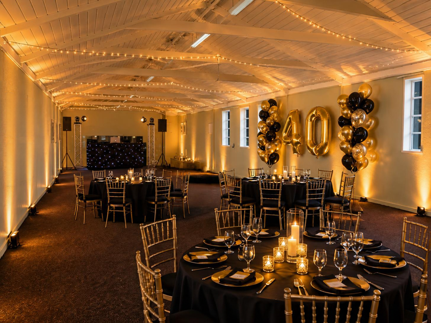 Elegant banquet hall with gold drapes, string lights, black table settings, and large “40” balloons