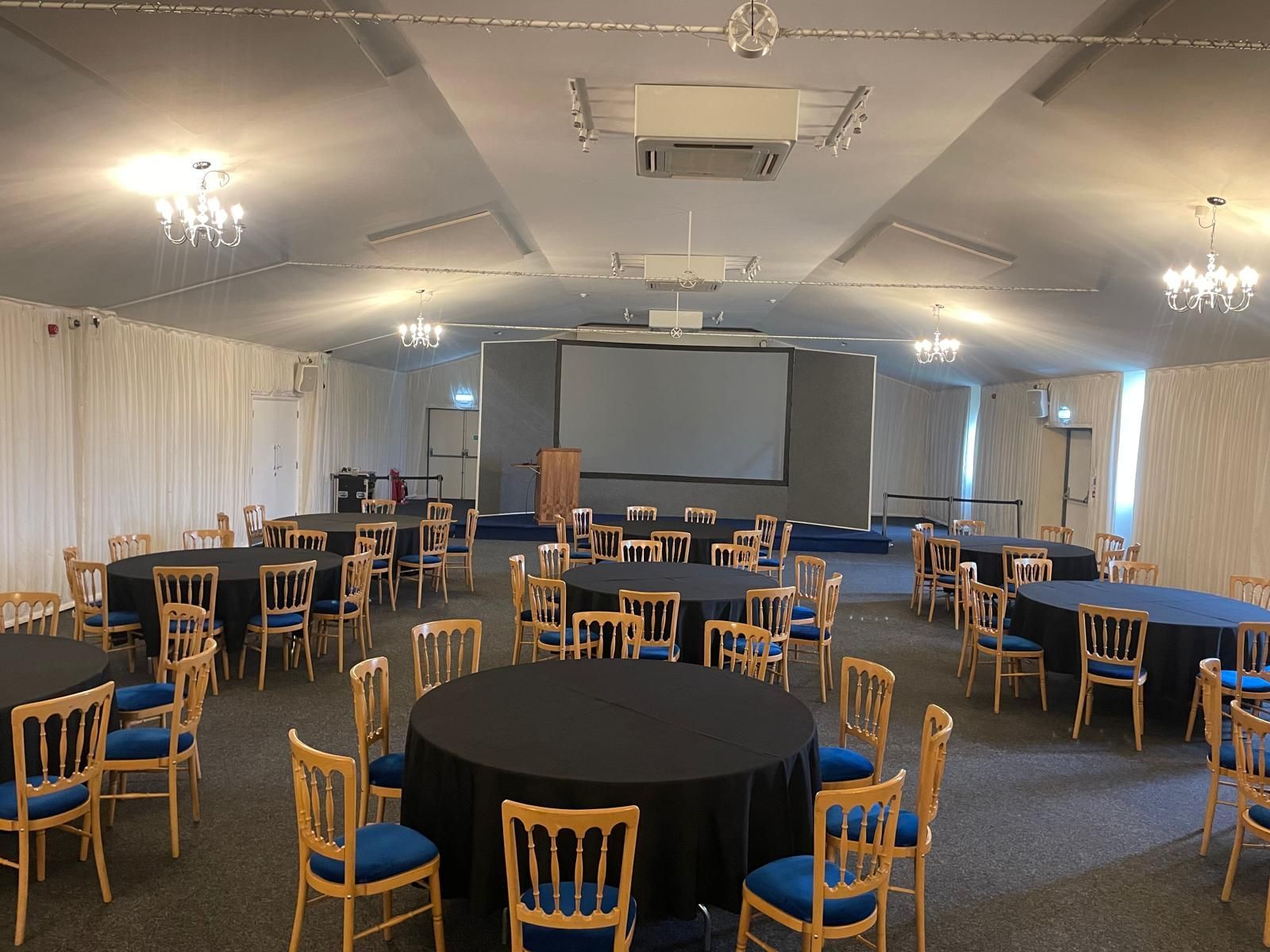 Empty banquet hall with round tables, blue chairs, and a projection screen at the front