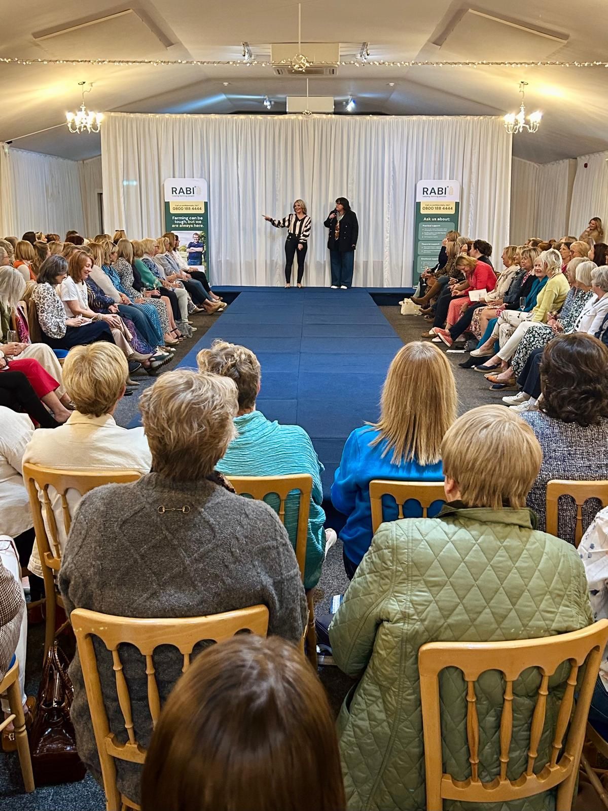 Audience seated in a tented hall, watching three speakers on a blue-carpeted stage.