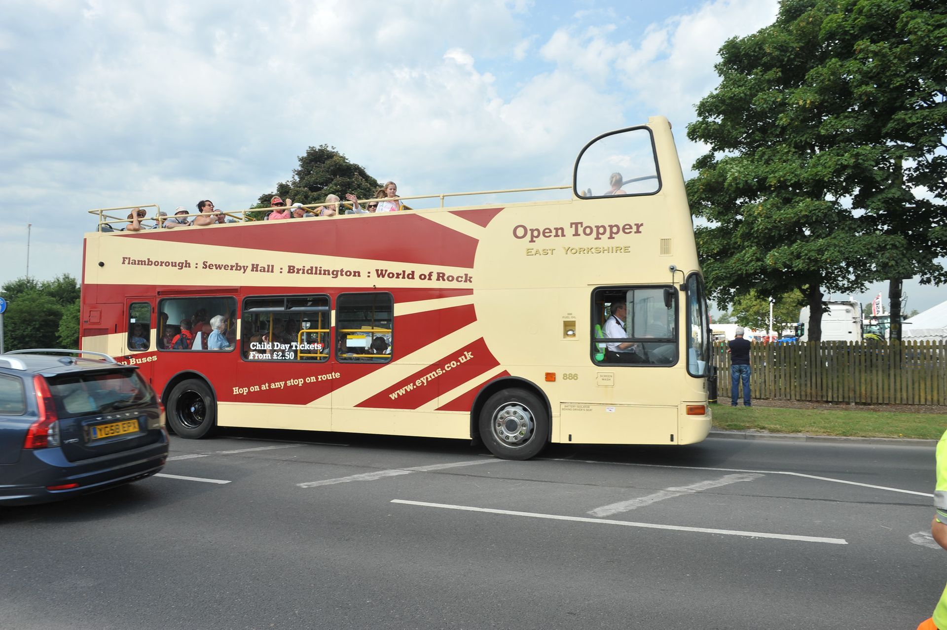 Double-decker tour bus on a road beside a parked car, with trees and a cloudy sky in the background