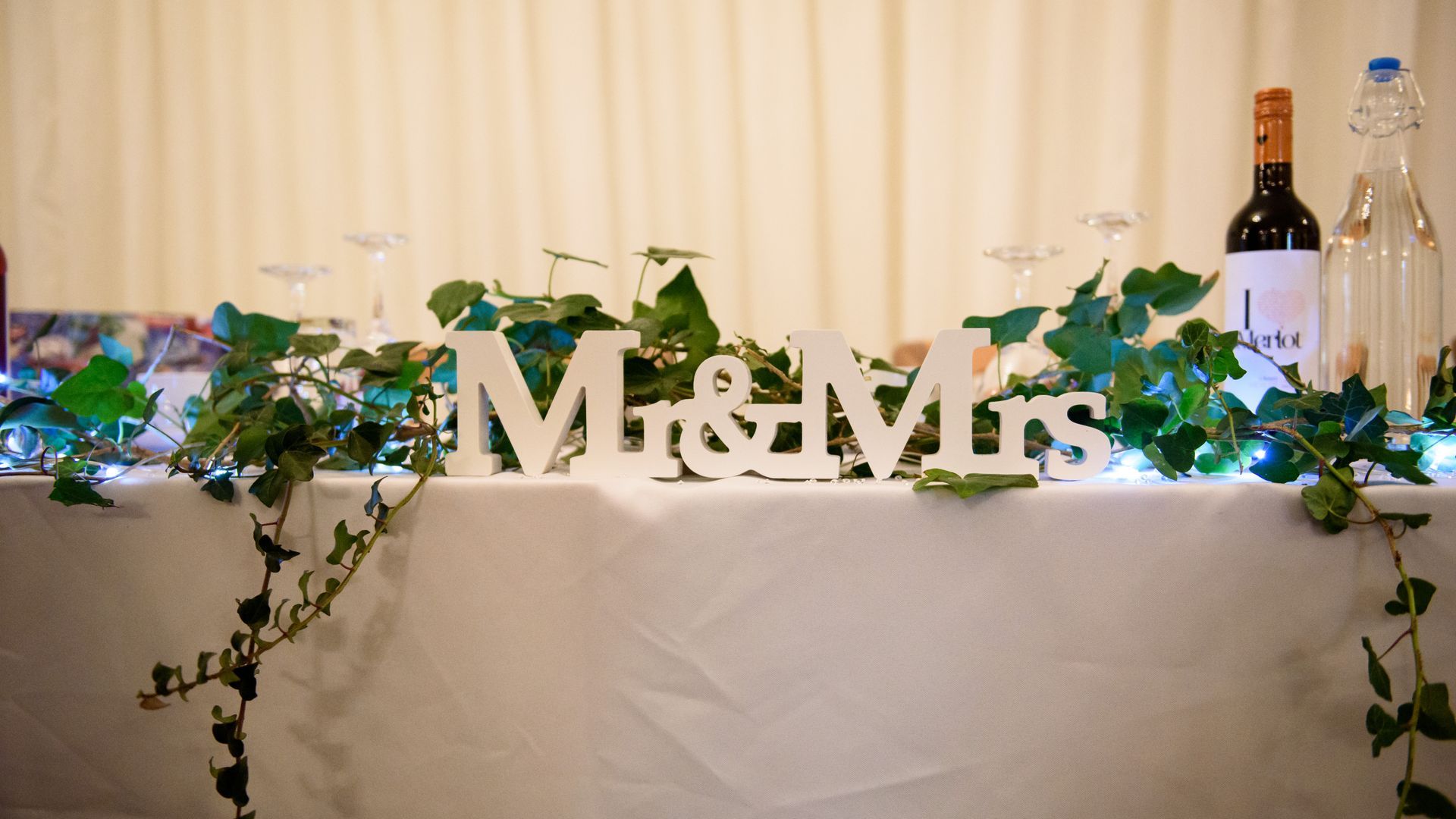 “Mr & Mrs” sign on a decorated wedding reception table with flowers, greenery, bottles, and a white tablecloth