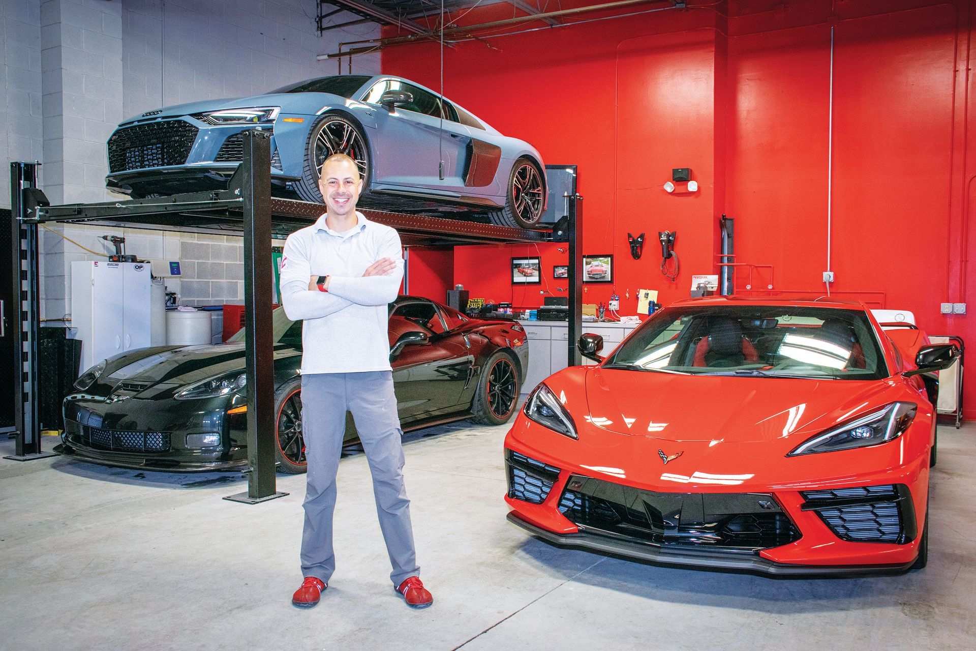 A person standing with arms crossed in a garage with a lifted grey Audi R8, a dark Corvette, and a red Corvette C8.