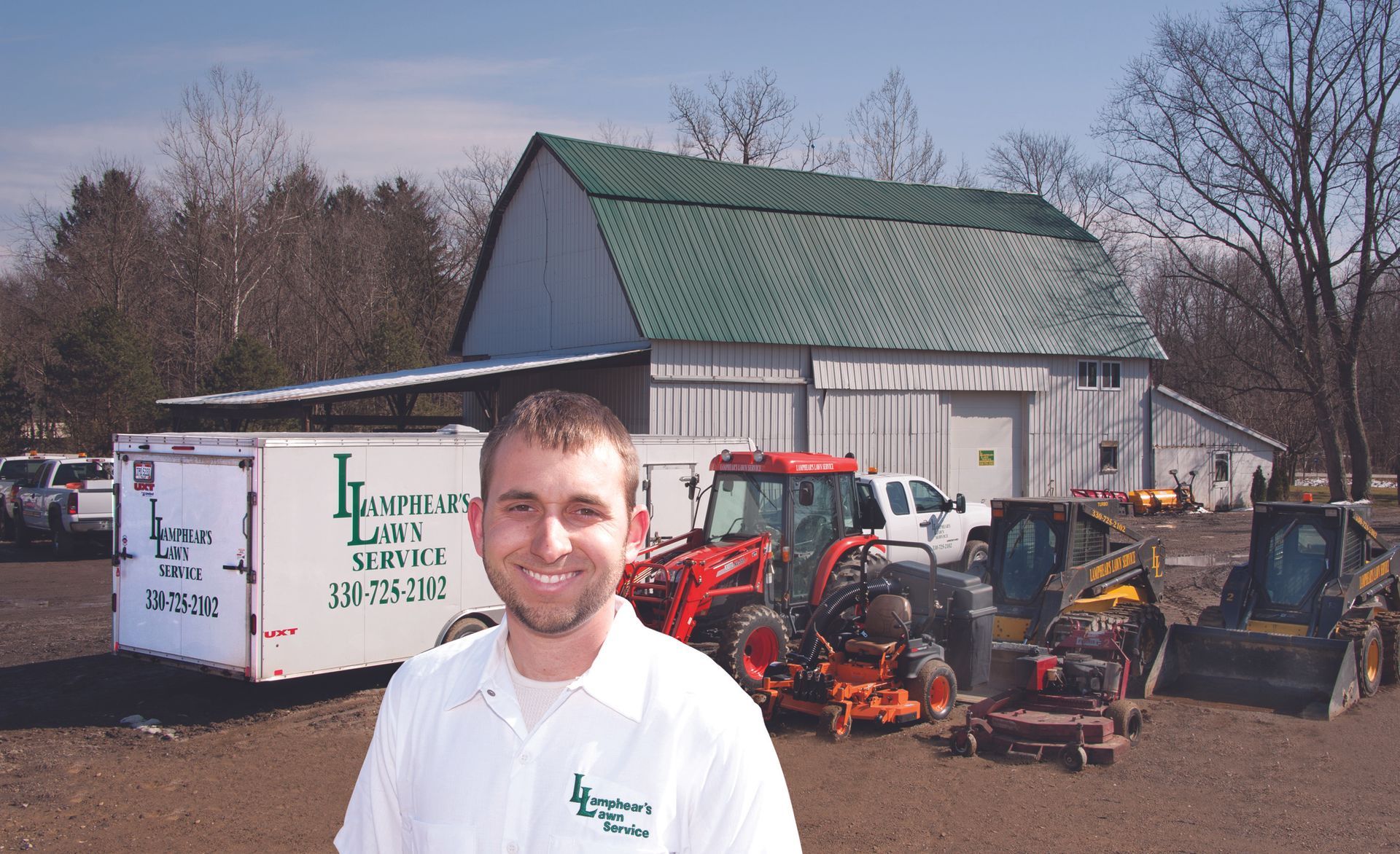 A smiling man in a white polo shirt stands in front of a barn, a white service trailer, and landscaping equipment.
