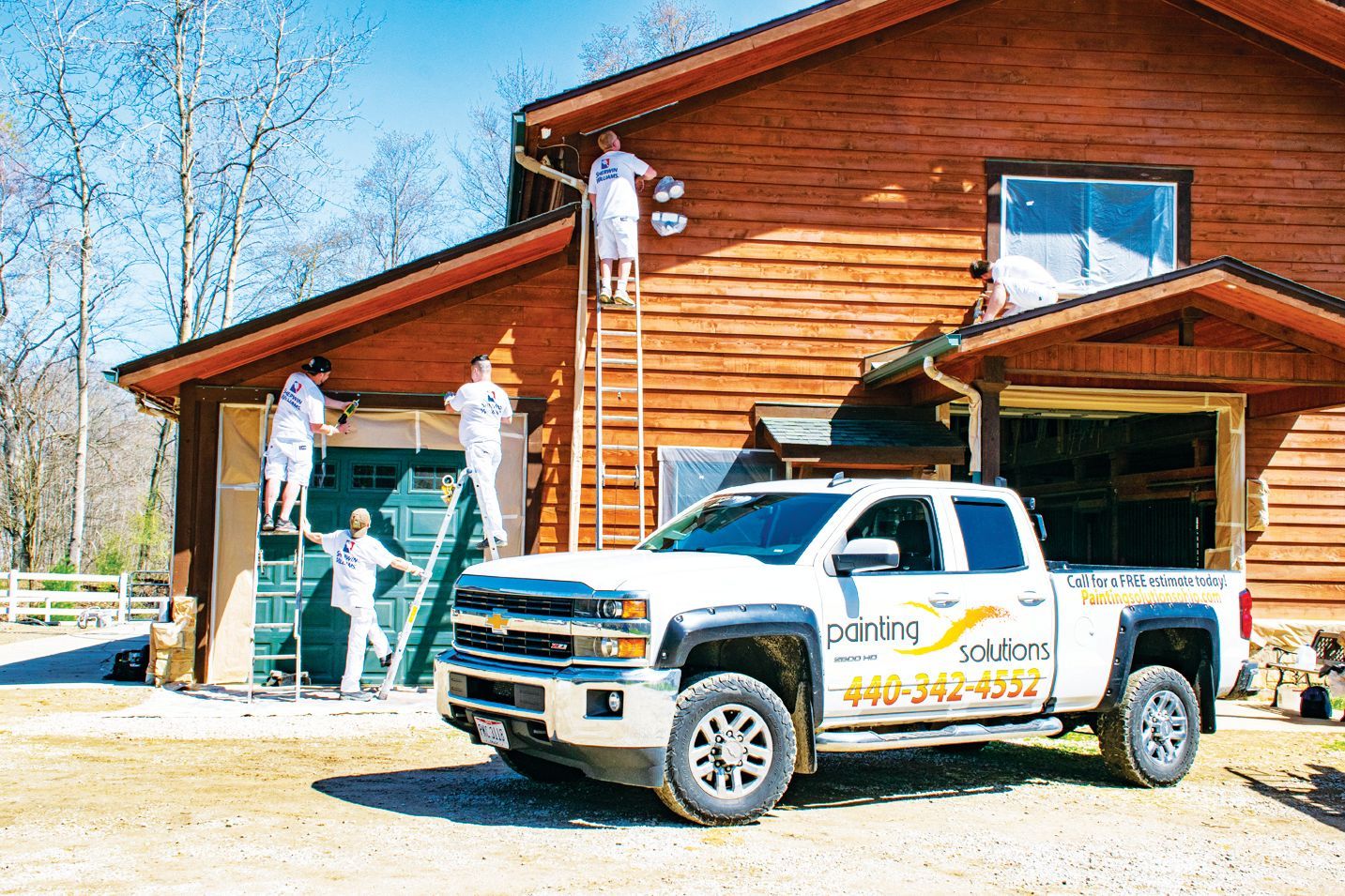 Four painters in white uniforms work on the exterior of a log building, with a company truck parked in the foreground.