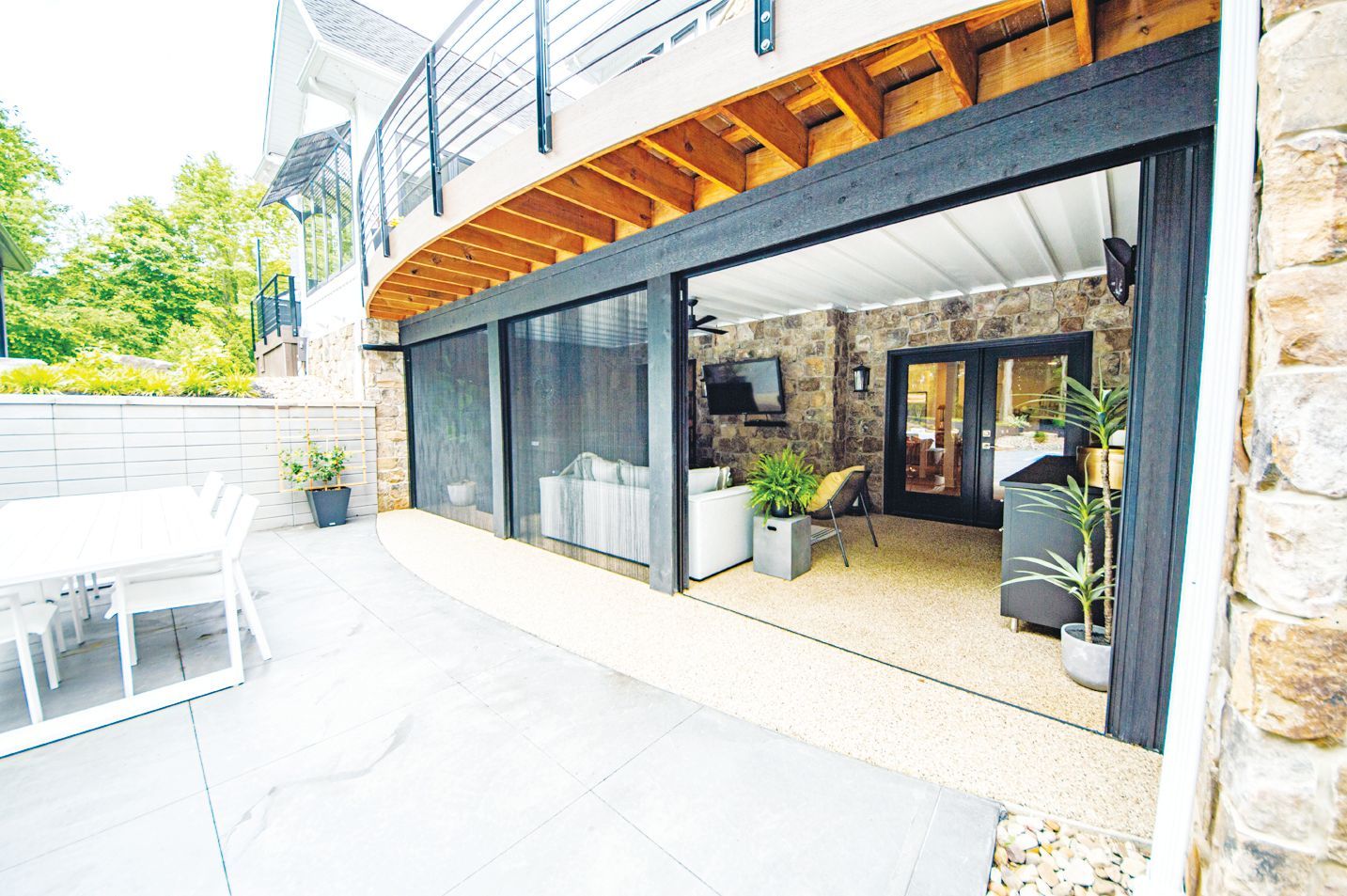 A patio features a white dining set on a concrete floor next to an open-air living area with a stone wall and sofa.