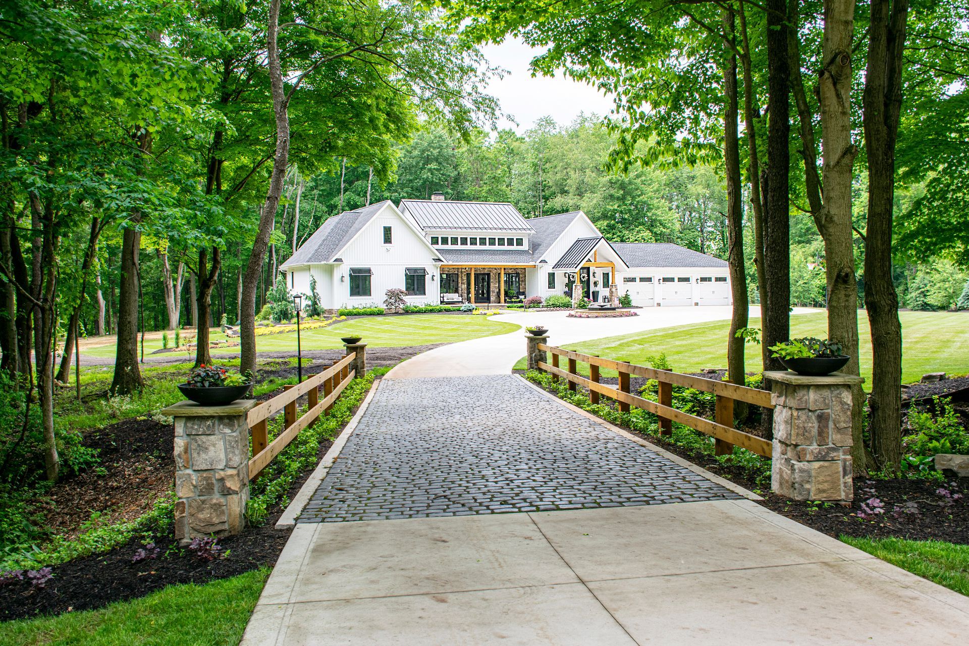 A white house with a multi-car garage, accessed by a cobblestone and concrete driveway lined by stone pillars and fences.