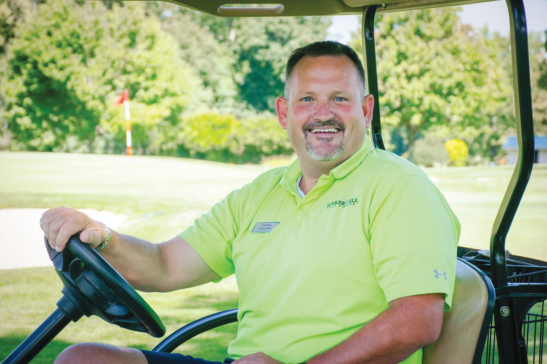 A smiling person wearing a lime green polo shirt sitting in the driver’s seat of a golf cart on a sunny golf course.
