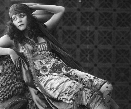 A person in an ornate, floral-patterned outfit and headwrap reclines against a textured background, posing pensively.