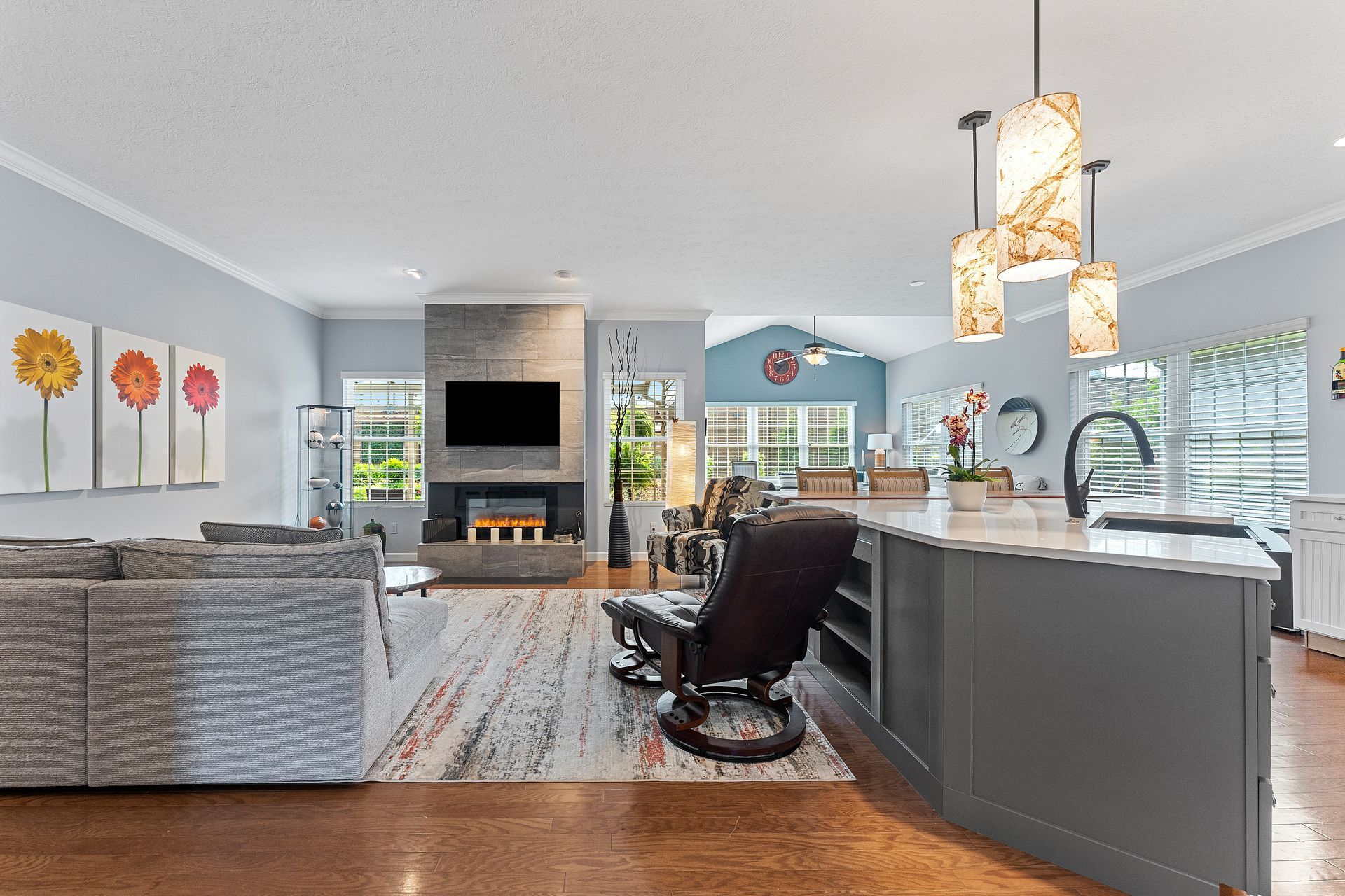 An open-concept living area with a grey sofa, fireplace, patterned rug, kitchen island, and three hanging light fixtures.