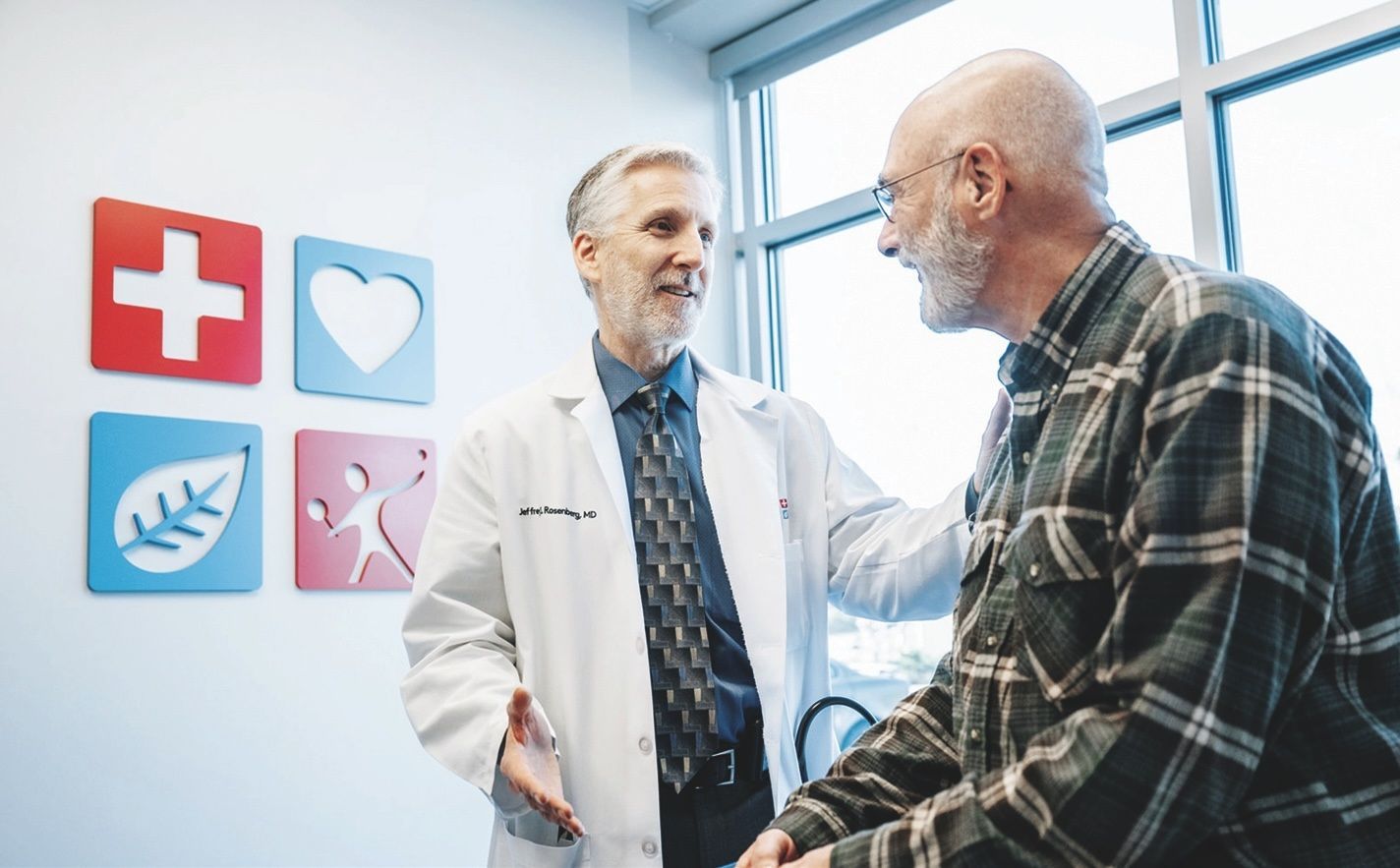 A doctor in a white coat talks with a patient in an office with colorful wall icons.
