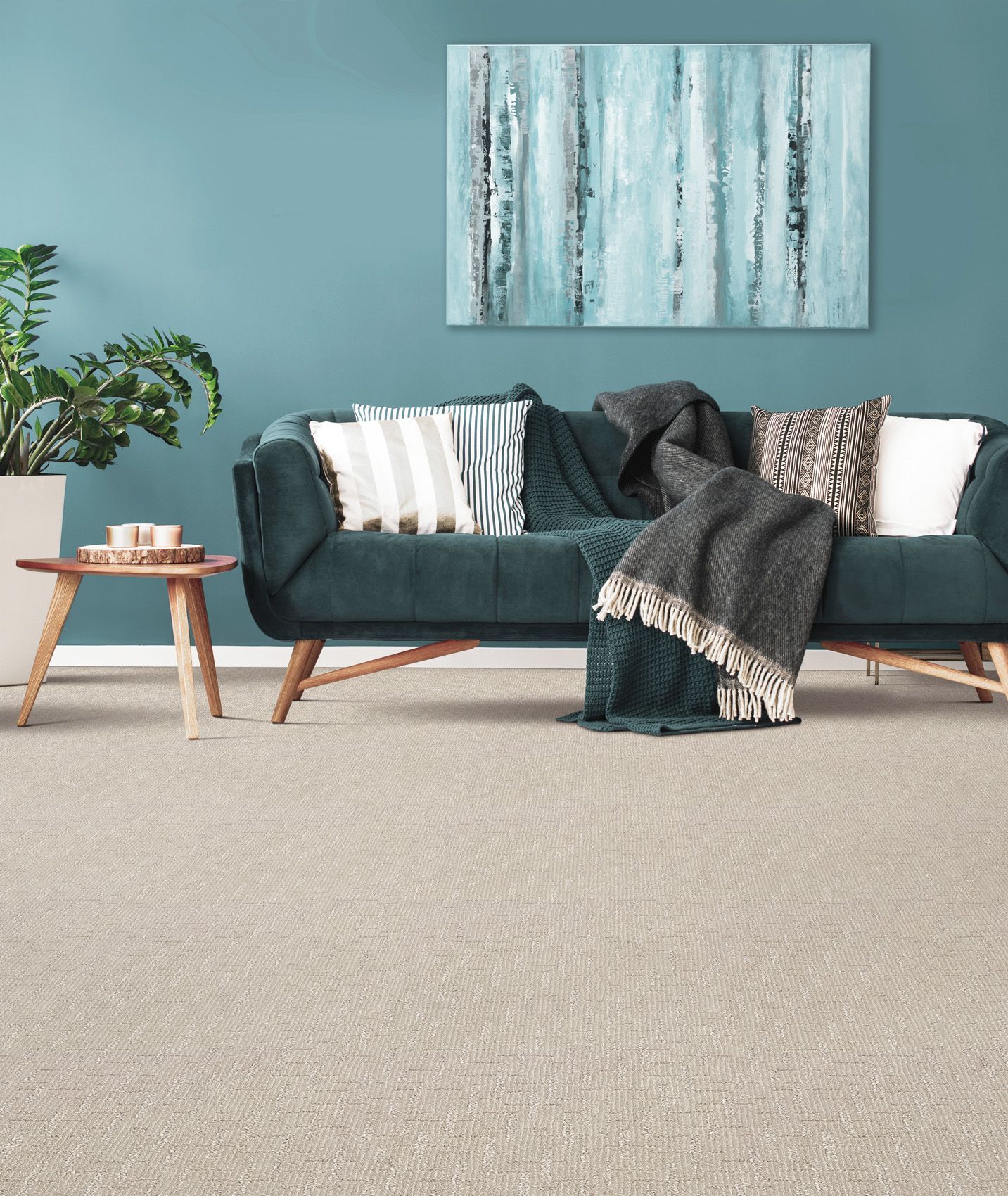 A teal sofa with patterned pillows and a dark throw blanket sits on a light carpeted floor against a blue wall with art.