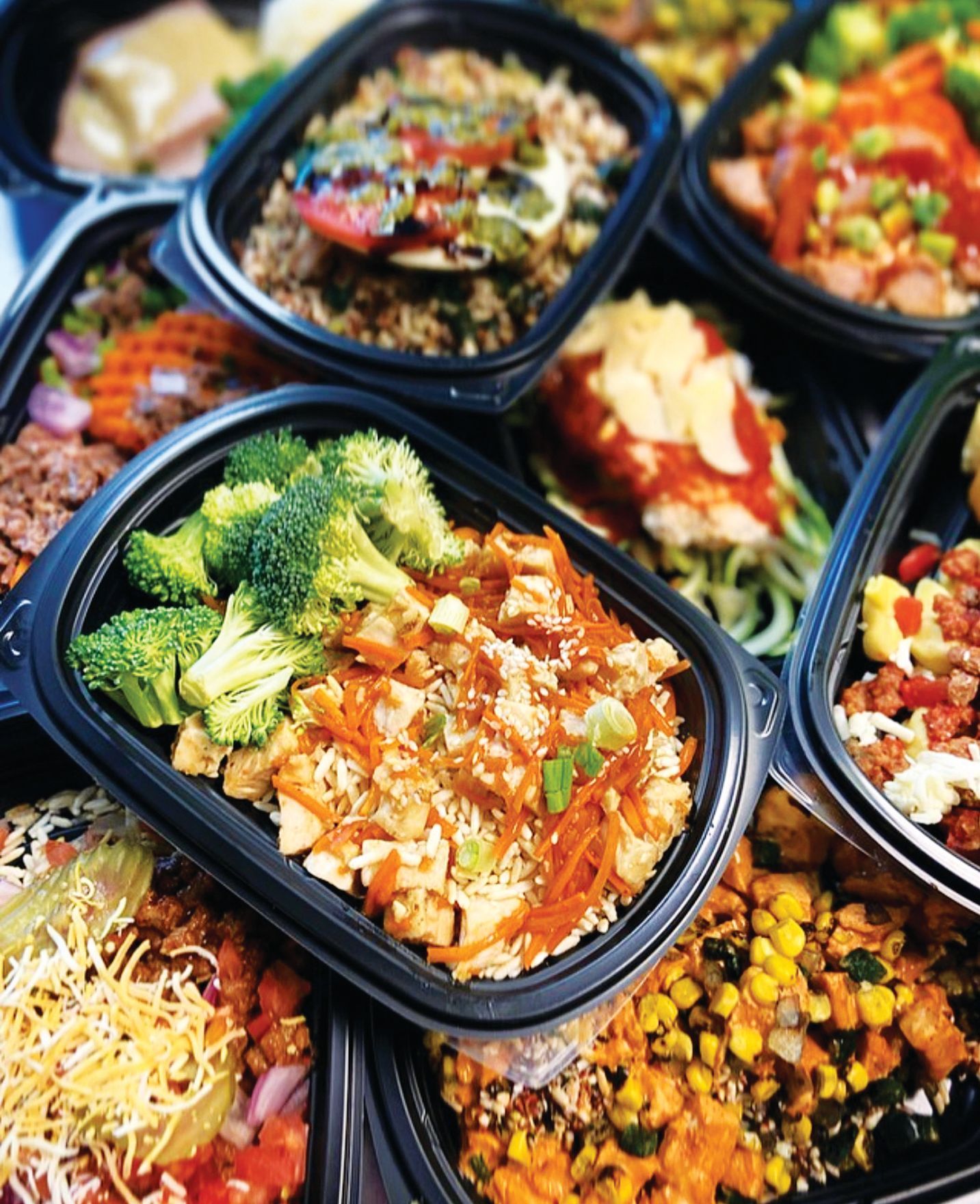A high-angle close-up of several black plastic containers filled with various colorful, prepared meal-prep dishes.