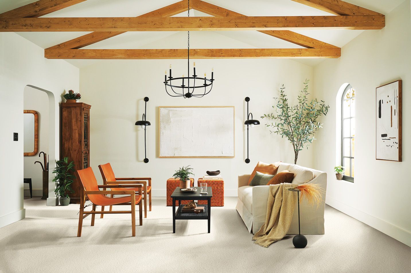 A bright living room with white walls, exposed wooden ceiling beams, two orange chairs, a beige sofa, and a chandelier.
