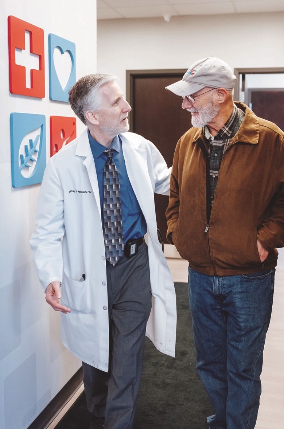 A clinician in a white lab coat walks and talks with a patient in a hallway decorated with colorful medical icons.