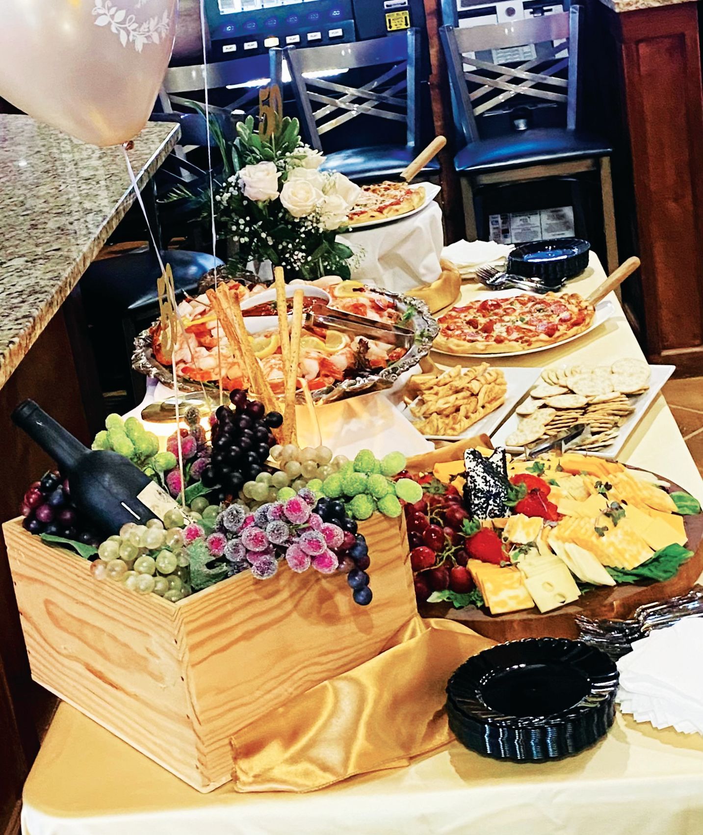 A spread of food including pizzas, crackers, cheese, and a wooden crate filled with assorted grapes and a wine bottle.