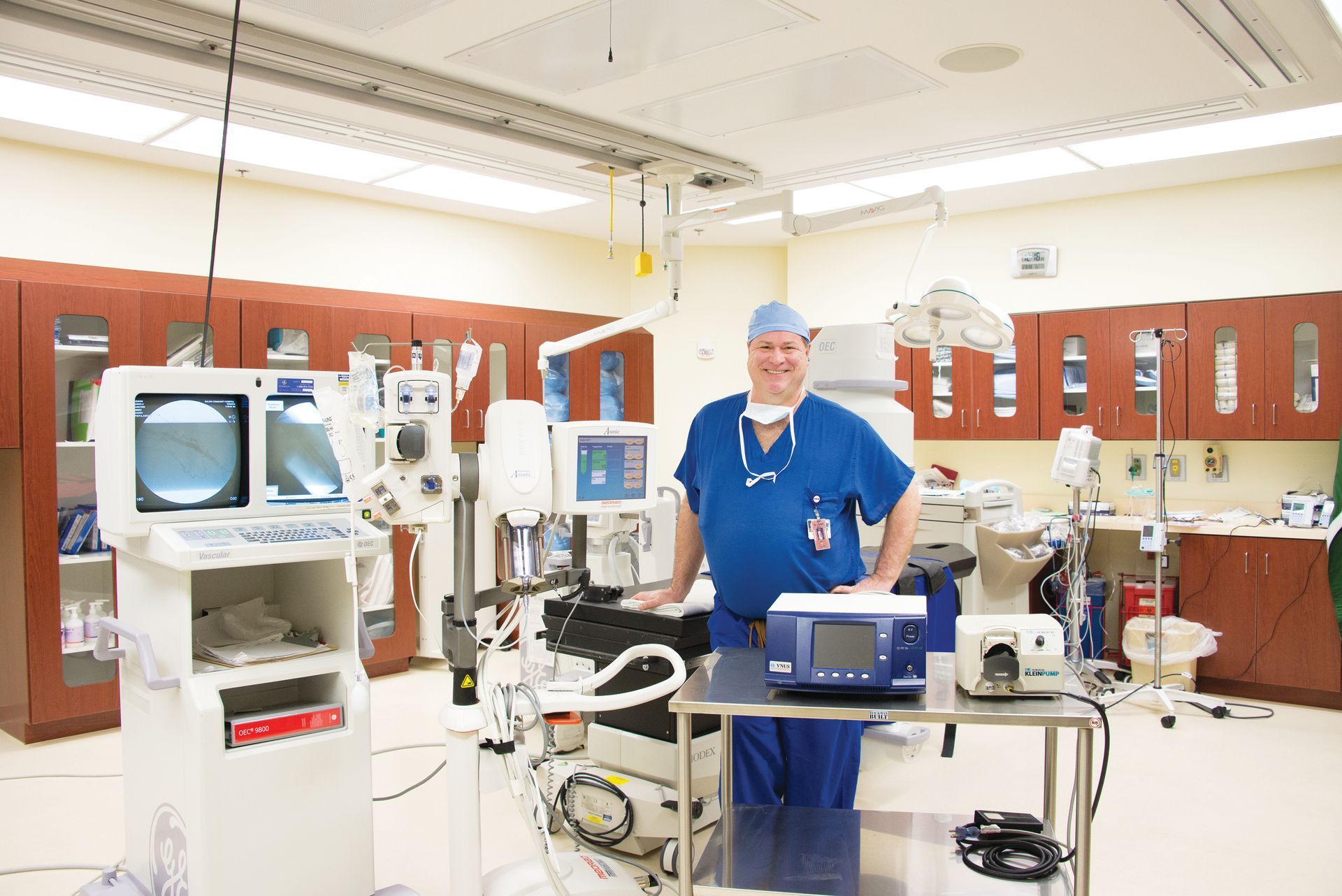 A person in blue surgical scrubs stands in a sterile operating room filled with medical equipment and diagnostic screens.
